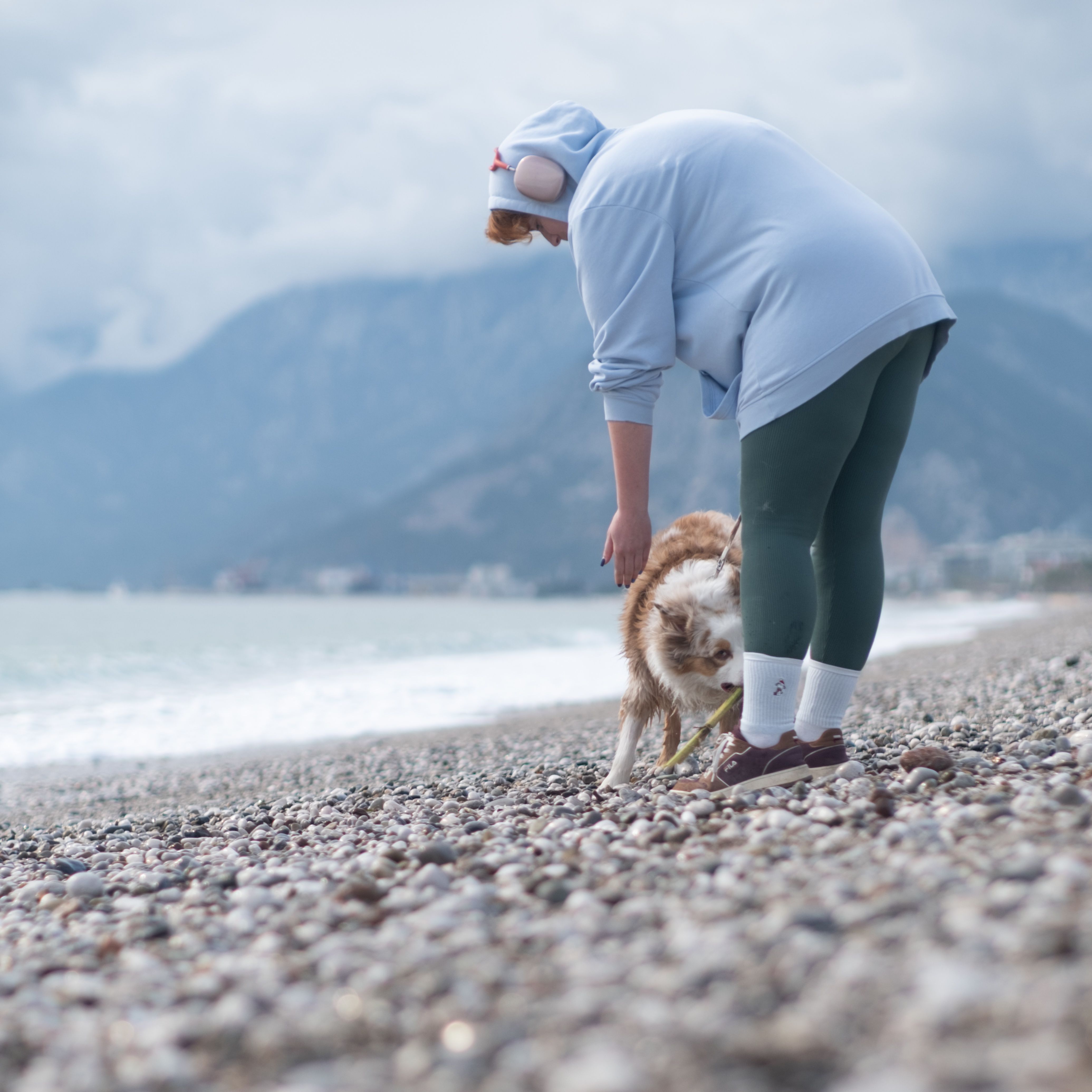 A woman and her dog enjoy a playful moment on a rocky beach with mountains in the background.