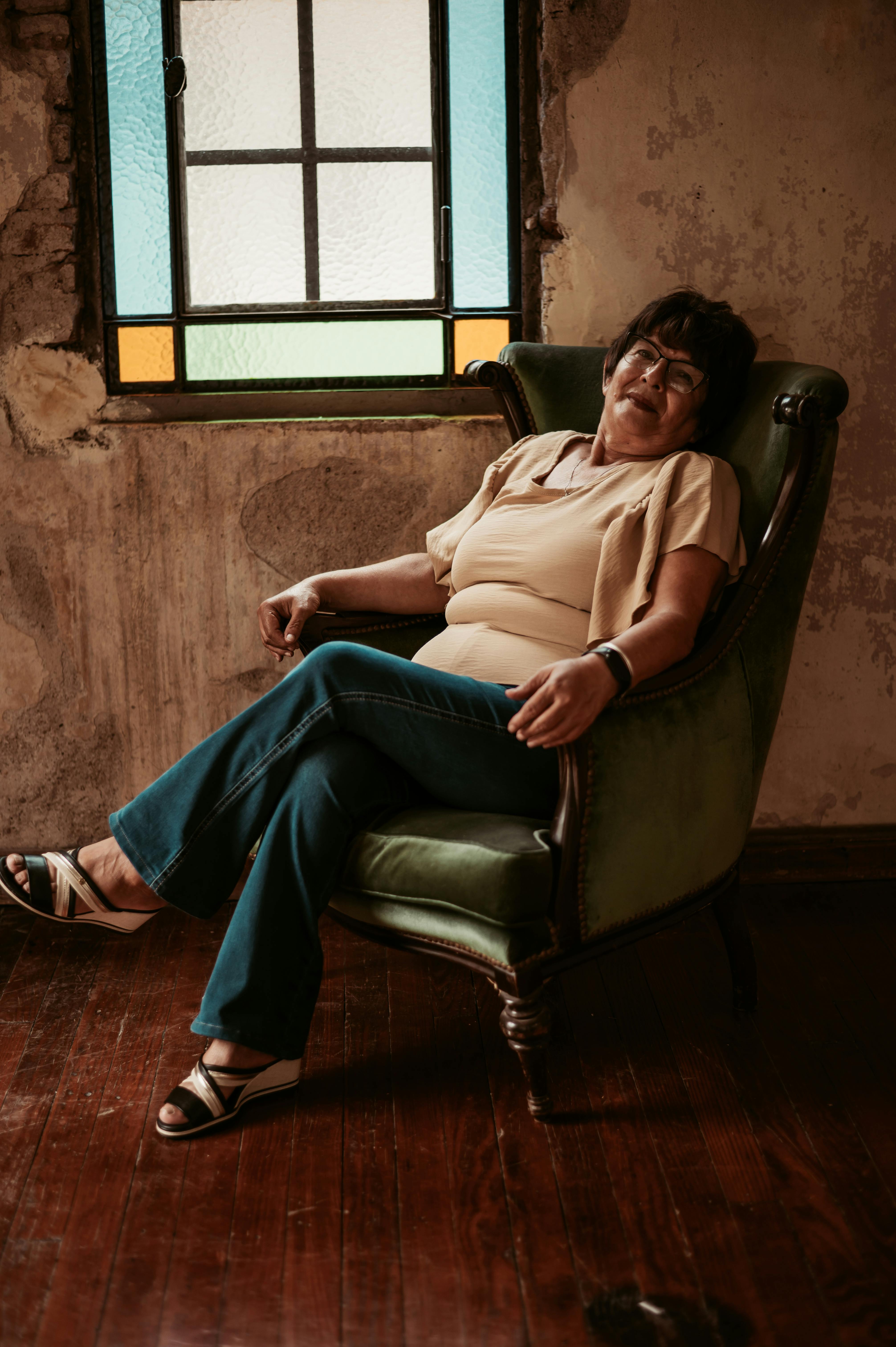 Безкоштовне Задоволена жінка розслабляється у старовинному кріслі біля барвистого вітража. стокове фото