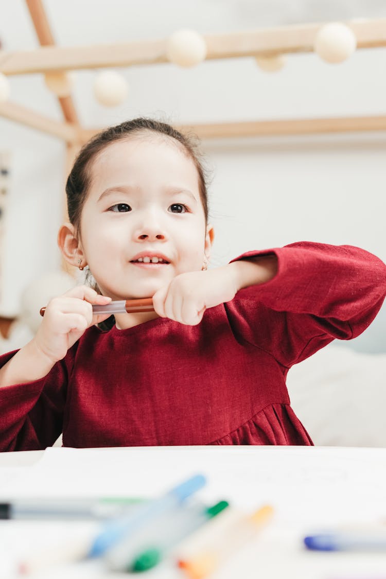 Selective Focus Photo Of Young Girl In Red Dress Holding Marker Pen While Seated At A Table