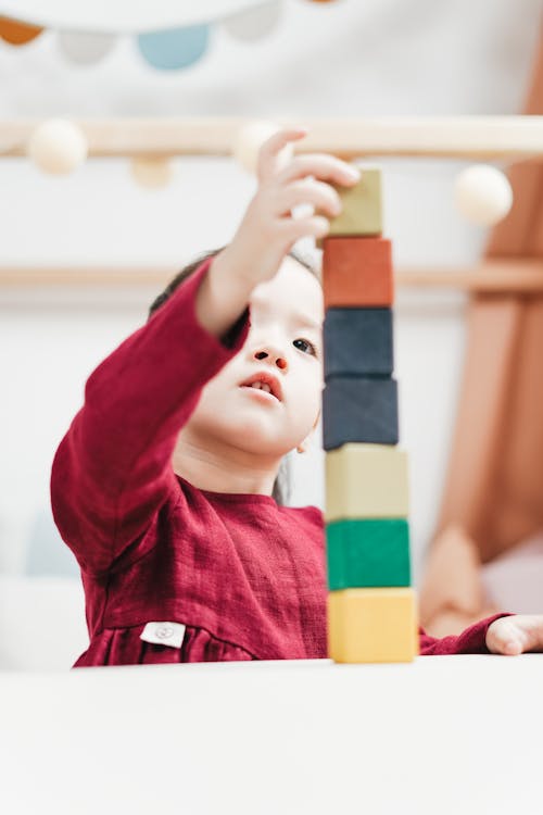 Selective Focus Photo of Young Girl in Red Dress Playing with Building Blocks