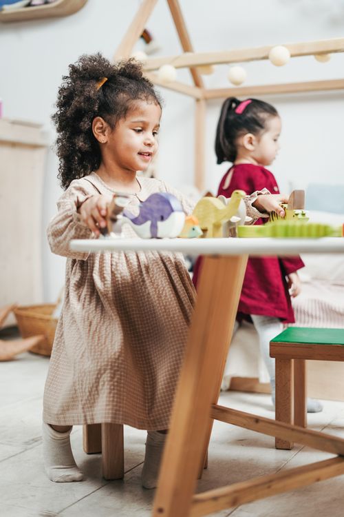 Two young girls playing with wooden toys and furniture in a bright playroom
