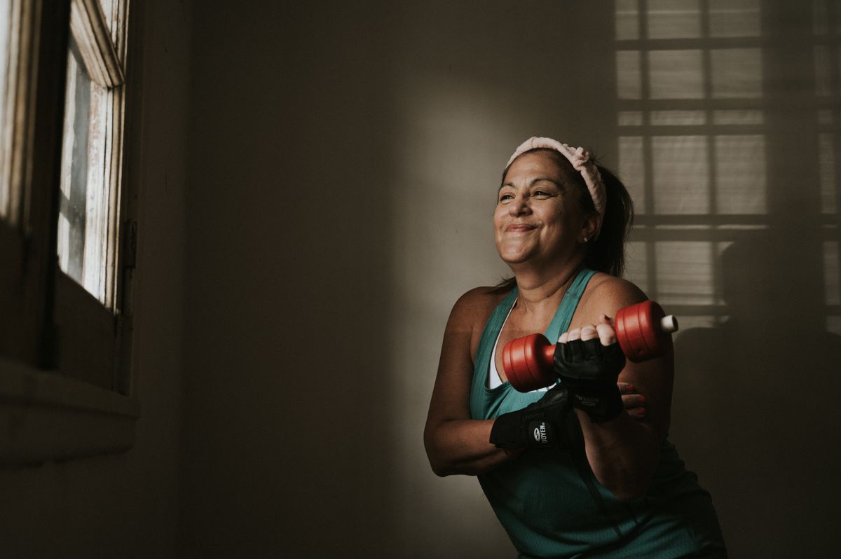 A middle-aged woman in a teal top smiling while lifting a red dumbbell indoors.