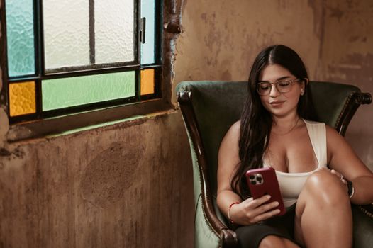 Young woman sitting in vintage chair, browsing smartphone inside a cozy, rustic room.