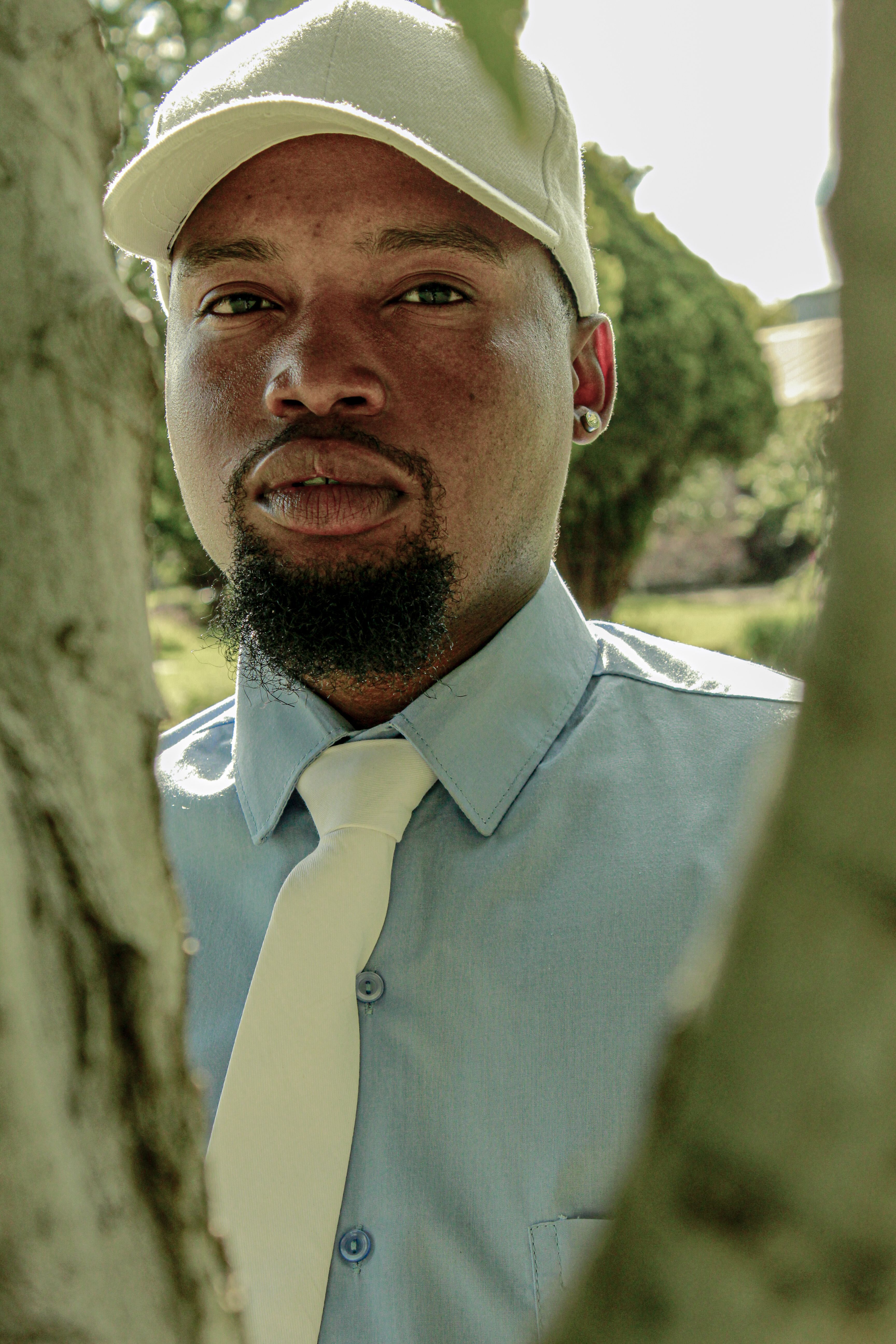 Close-up portrait of a man in a white cap and tie, outdoors surrounded by trees.