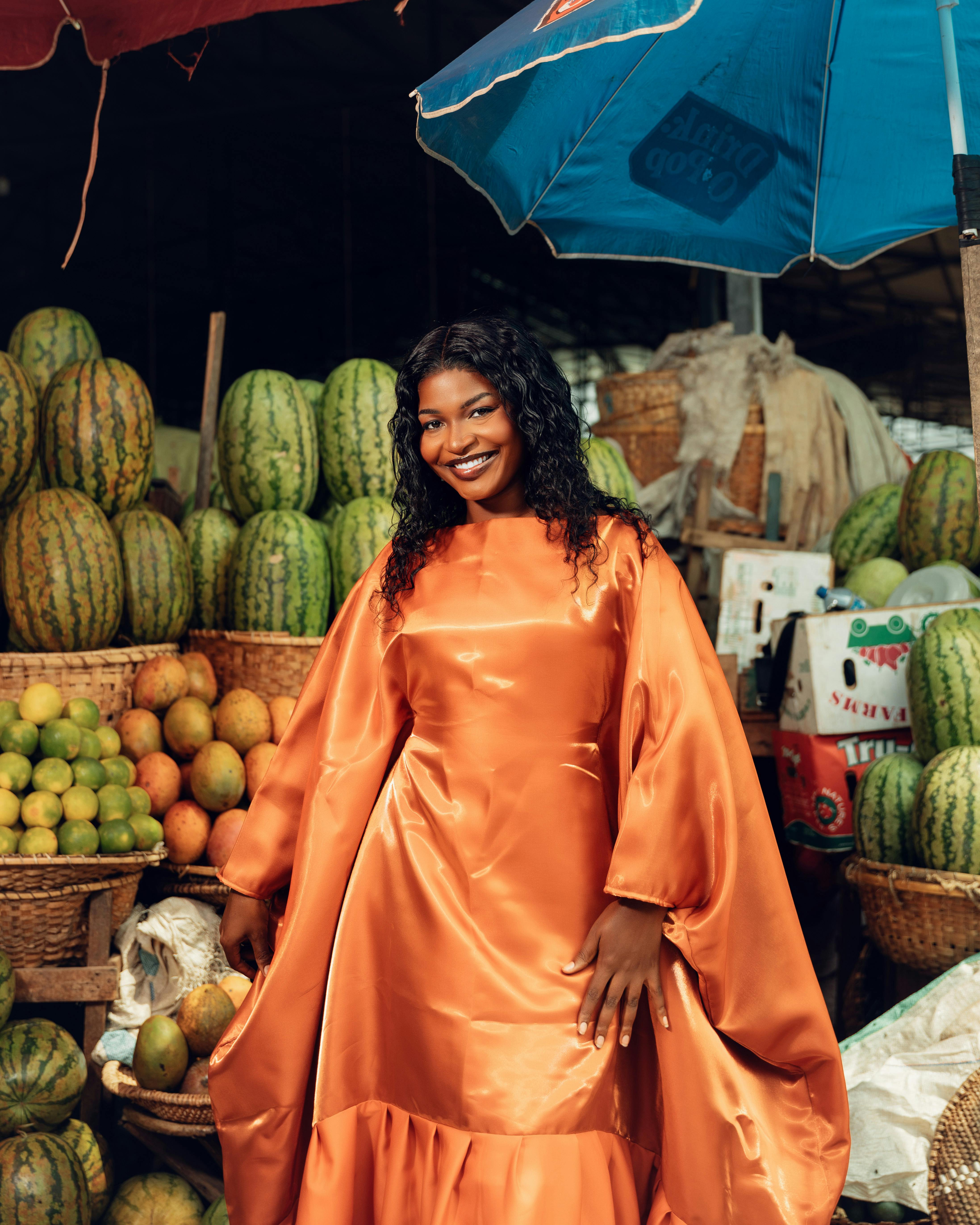 免費 一位身穿鮮豔橙色連身裙的微笑女子在色彩繽紛的水果攤前。 圖庫相片