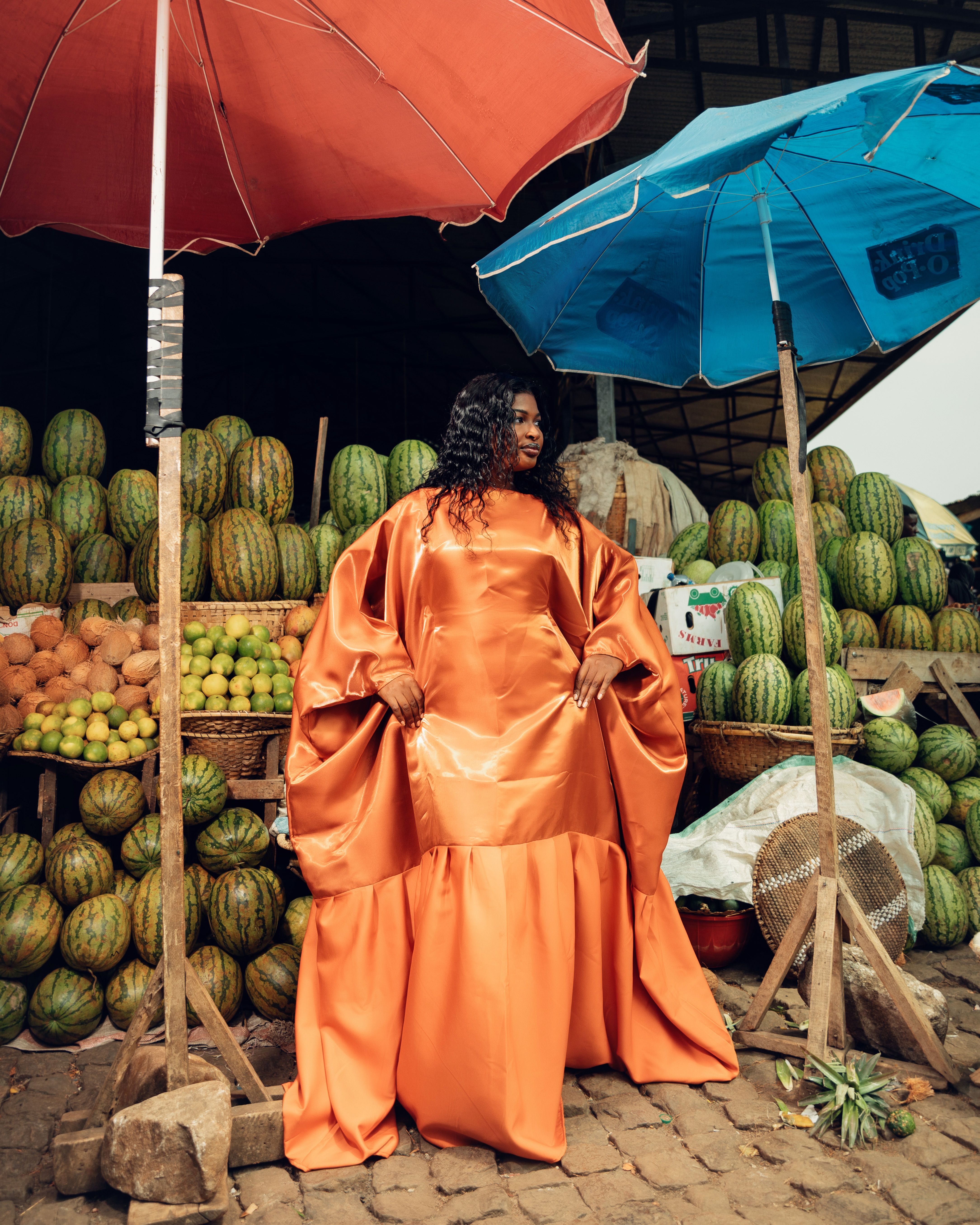 A stylish woman in a bold dress poses amidst colorful fruits at a market.