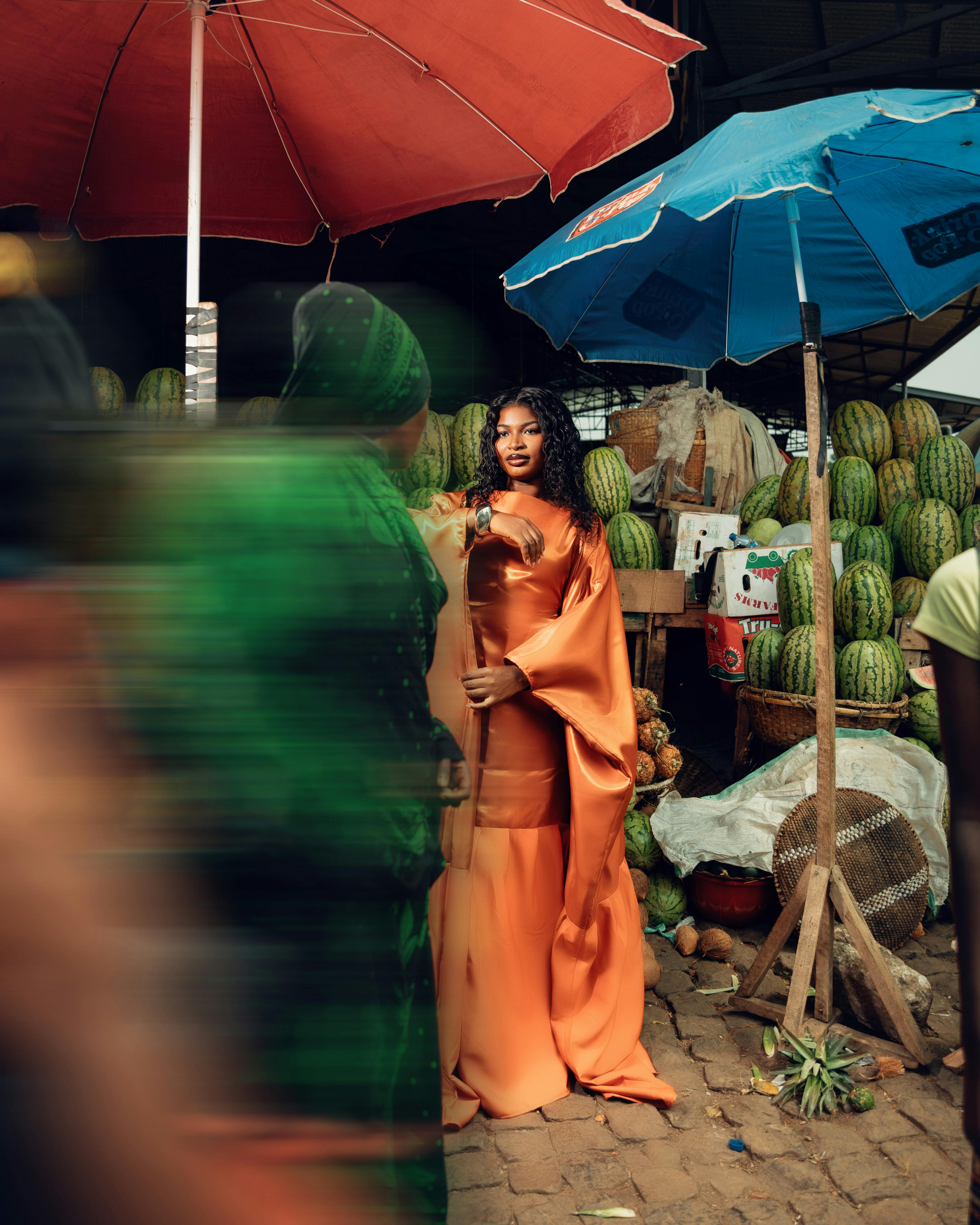 ฟรี หญิงสาวในชุดเดรสสีปะการังหรูหรา ยืนอยู่ท่ามกลางตลาดกลางแจ้งที่มีร่มกันแดดสีสันสดใส คลังภาพถ่าย