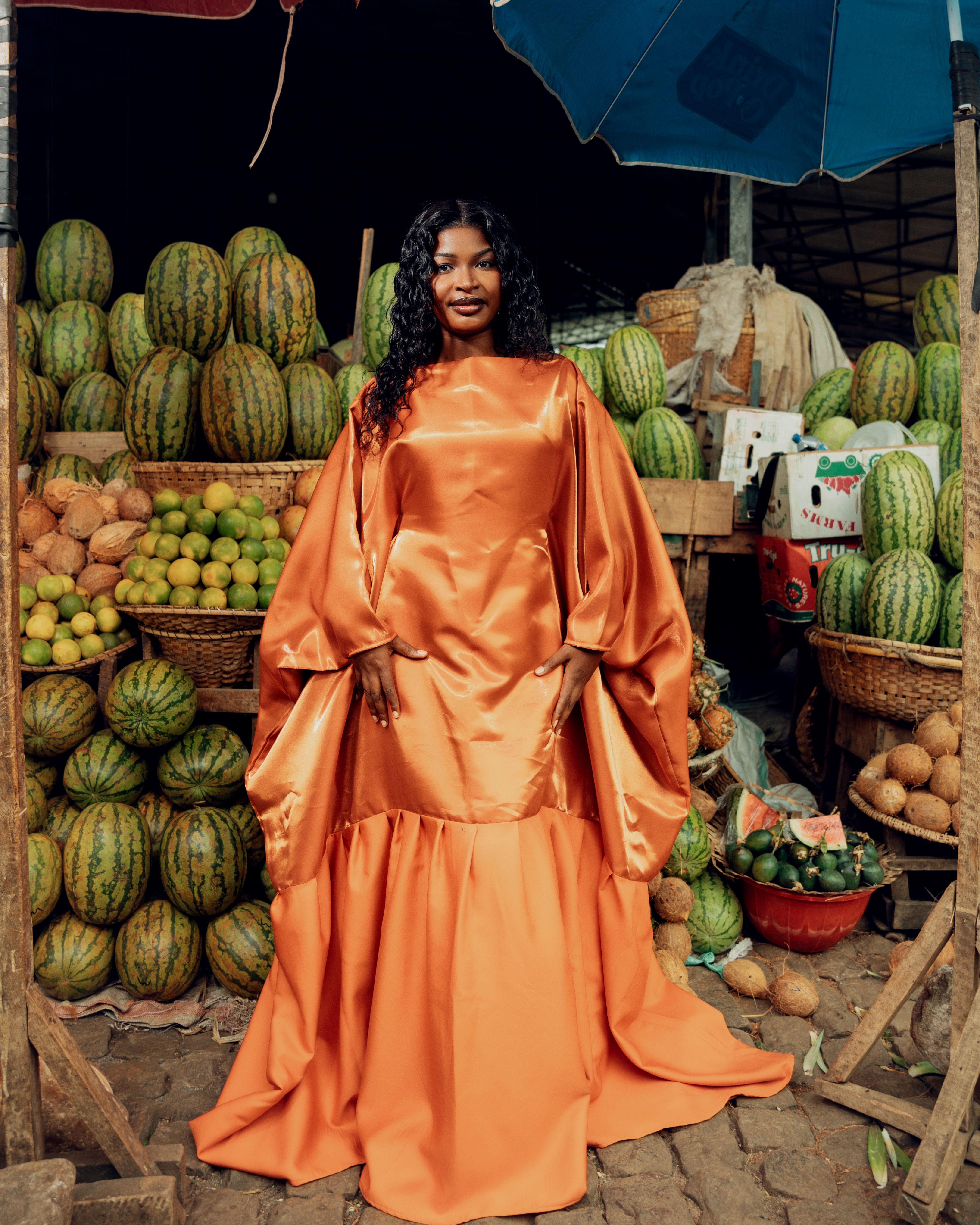 Fashionable woman in an orange gown poses in a colorful fruit market. Lively scene with melons and limes.