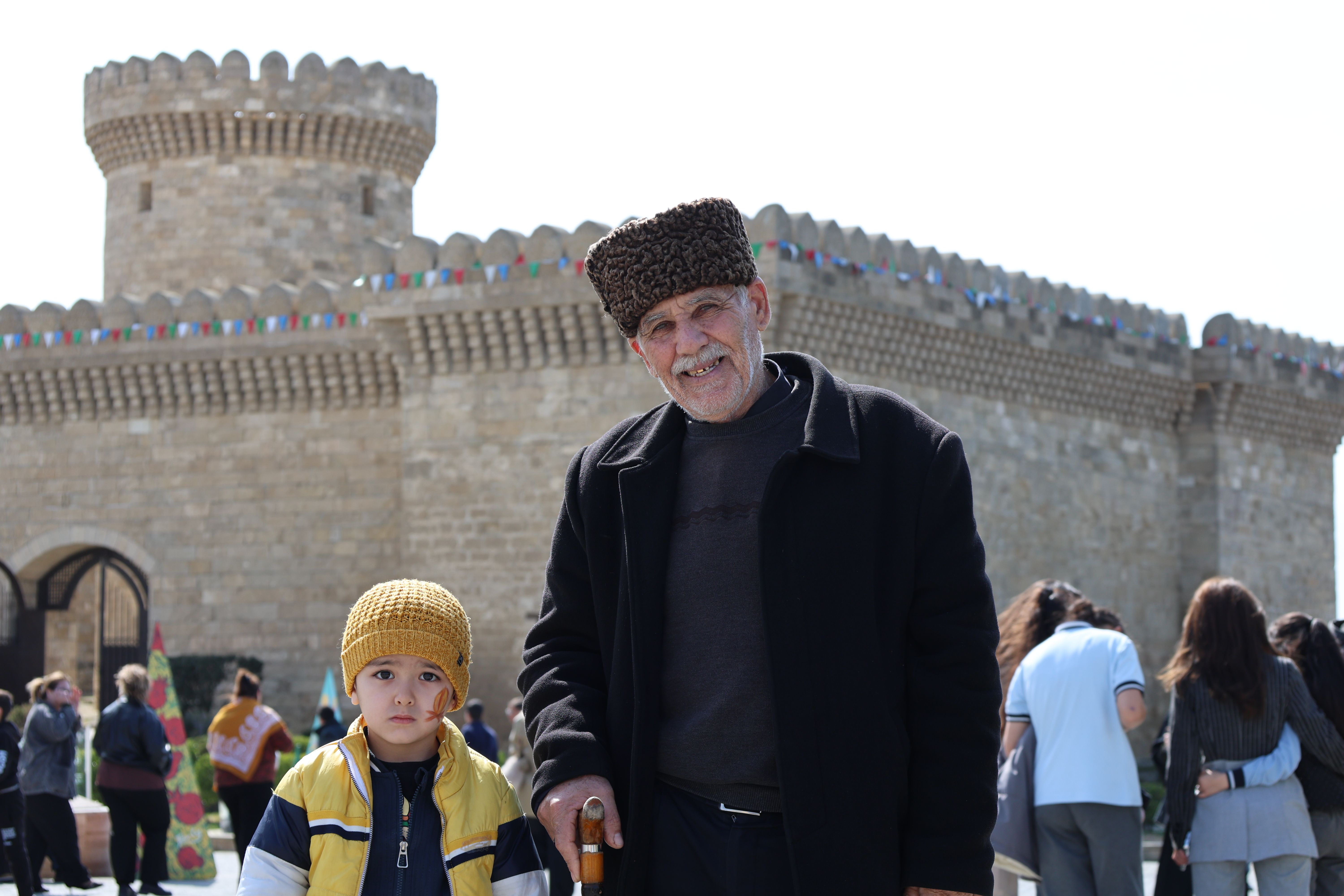 Free An elderly man with a cane and a child stand in front of a historic stone castle on a sunny day. Stock Photo