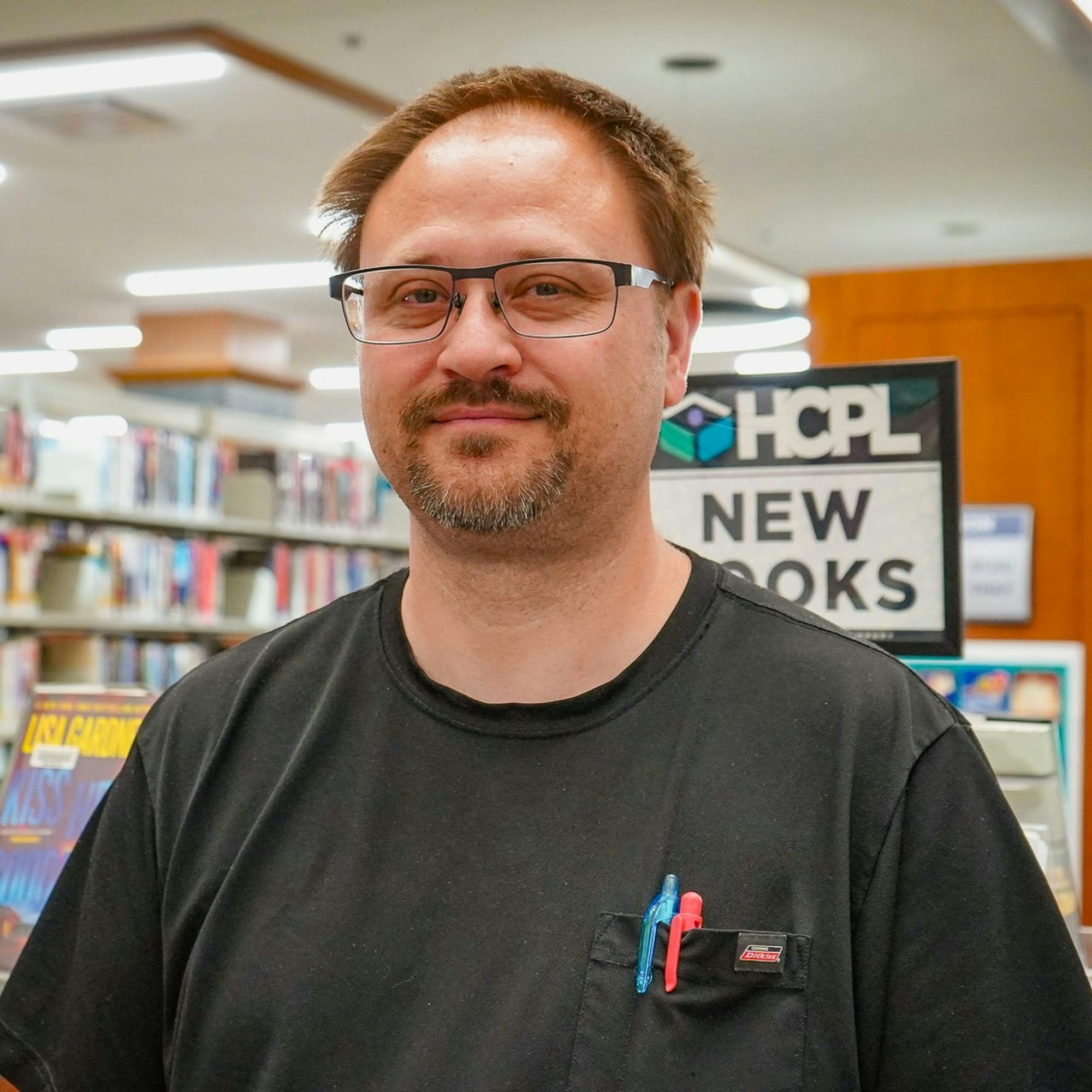 Man with glasses standing in a library near a 'new books' section.