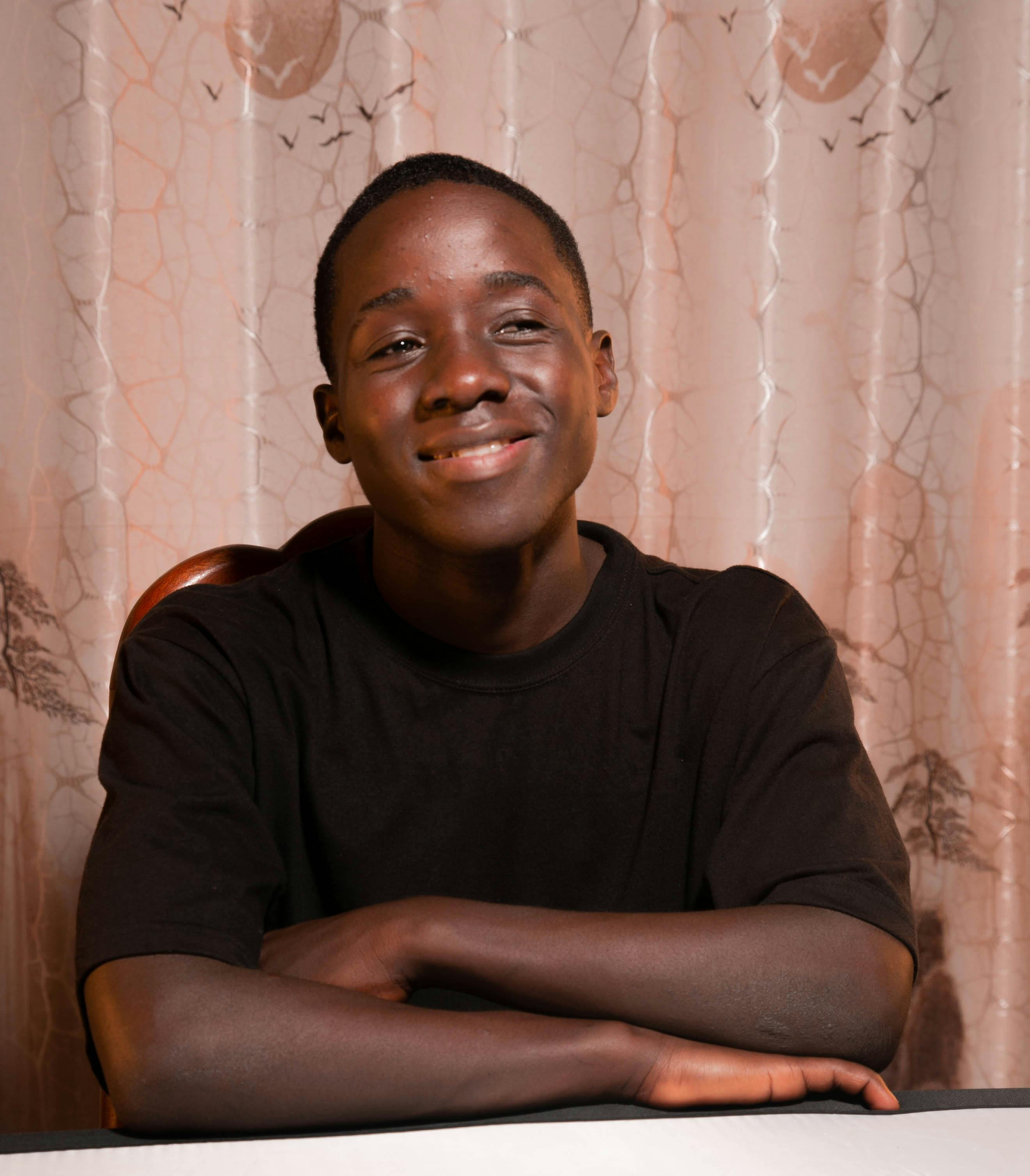 Portrait of a smiling teenage boy sitting indoors with arms crossed at a table.
