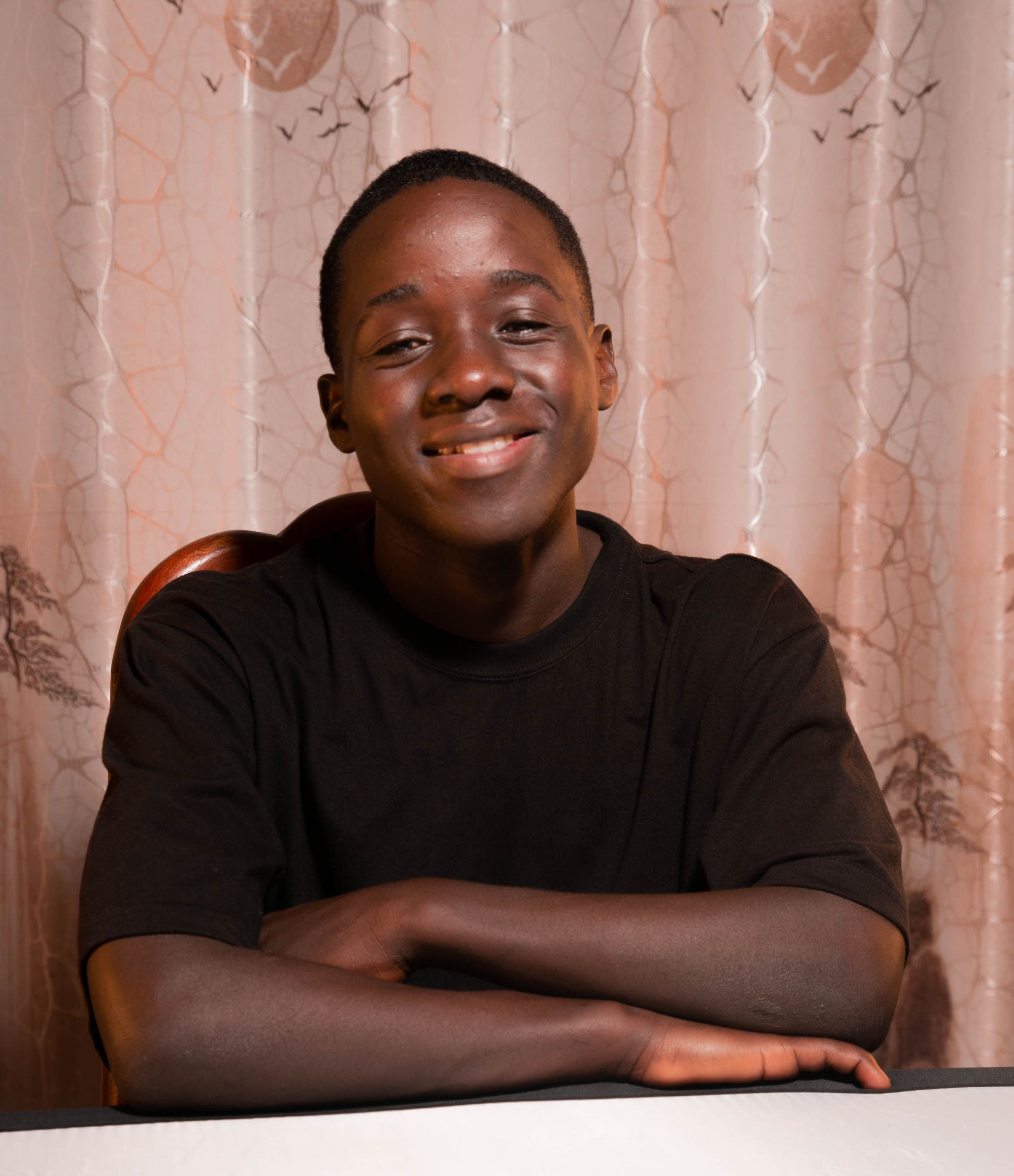 Portrait of a smiling teenager sitting at a table indoors with a curtain backdrop.