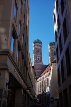 Frauenkirche towers seen between buildings in a Munich street on a clear day.