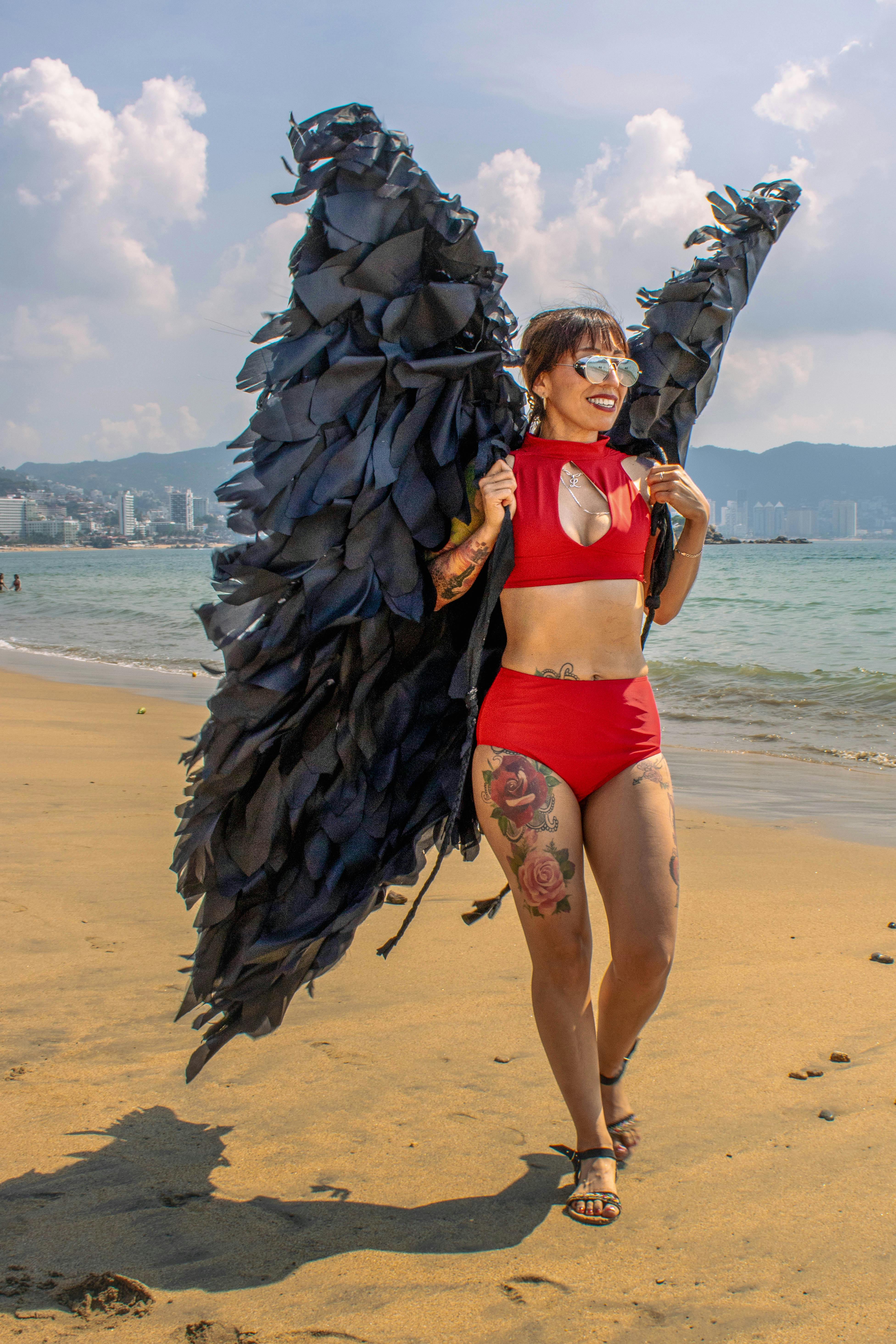 무료 아카풀코 해변에서 빨간색 수영복과 커다란 깃털 날개를 착용한 여성이 생동감 넘치는 해안 패션을 표현하고 있다. 스톡 사진