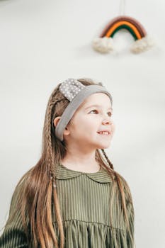 一个可爱的孩子穿着绿色连衣裙，扎着辫子，戴着头带，在彩虹墙装饰前微笑着