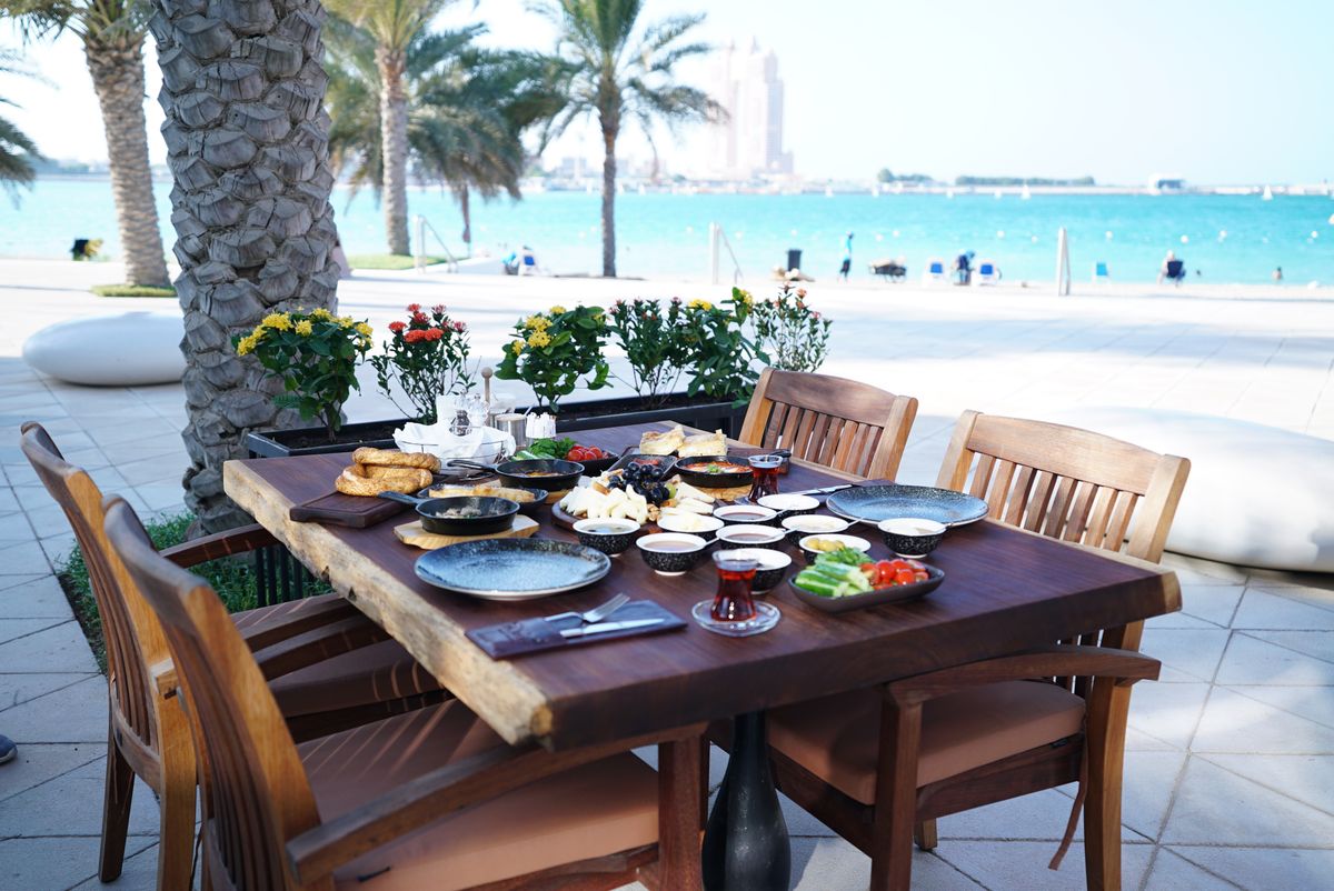 A long wooden table of mezze, breads and tea under palms beside the water