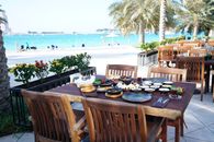 Elegant dining setup by a turquoise sea, surrounded by lush palm trees and vibrant flowers.