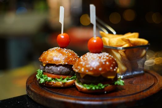 Gourmet mini burgers with cherry tomatoes and fries on a wooden platter in a restaurant setting.
