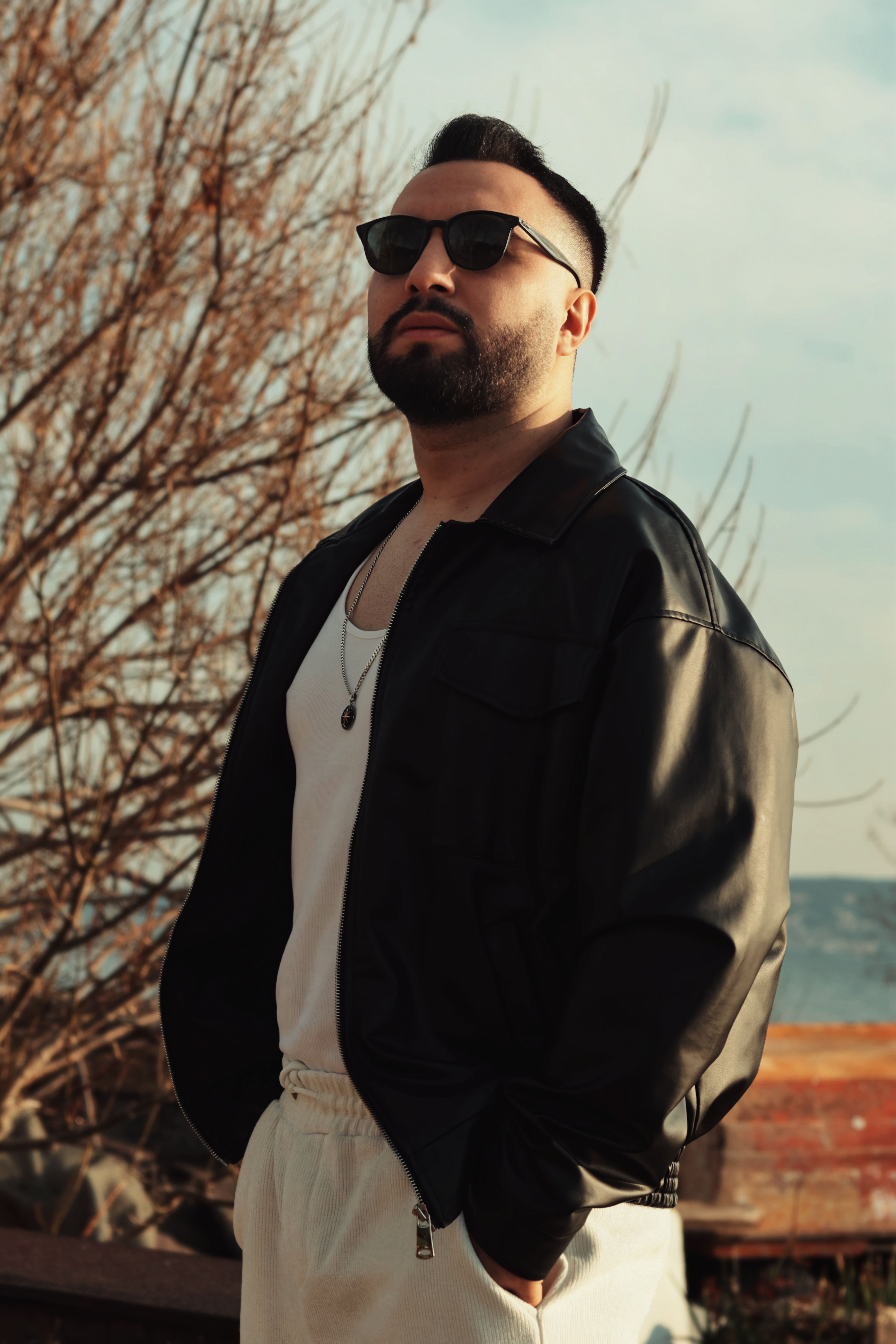 Confident man in sunglasses and jacket poses outdoors on a sunny day.