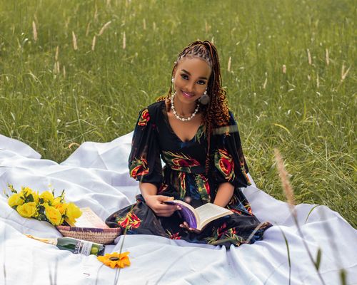 Free A woman in a floral dress reading a book while sitting on a picnic blanket in a grassy meadow. Stock Photo