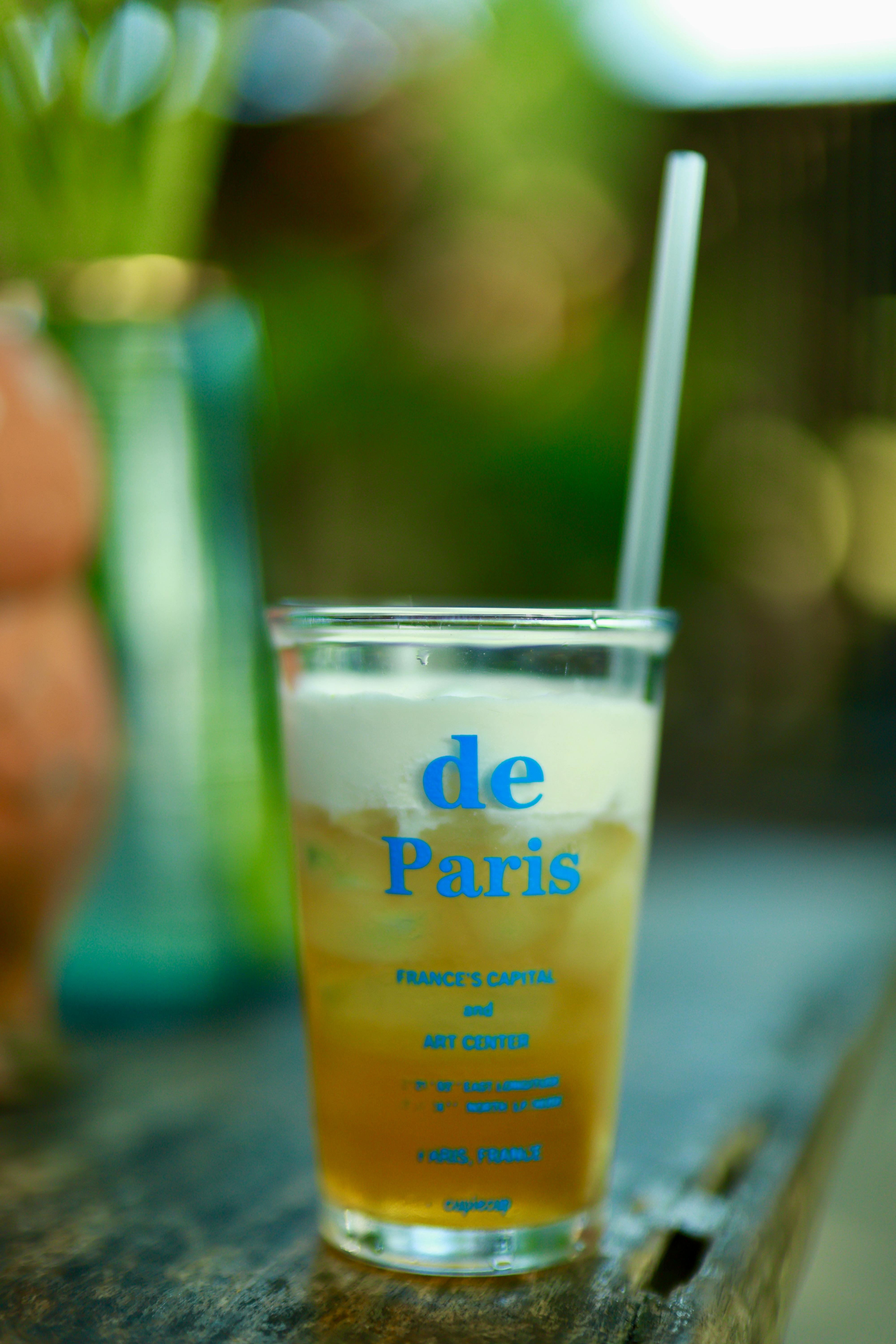 무료 빨대가 꽂힌 얼음 음료가 투명한 컵에 담겨 야외에 놓여 있고, 컵에는 'de Paris'라는 글자가 쓰여 있으며, 배경은 녹색으로 흐릿하게 처리되어 있다. 스톡 사진