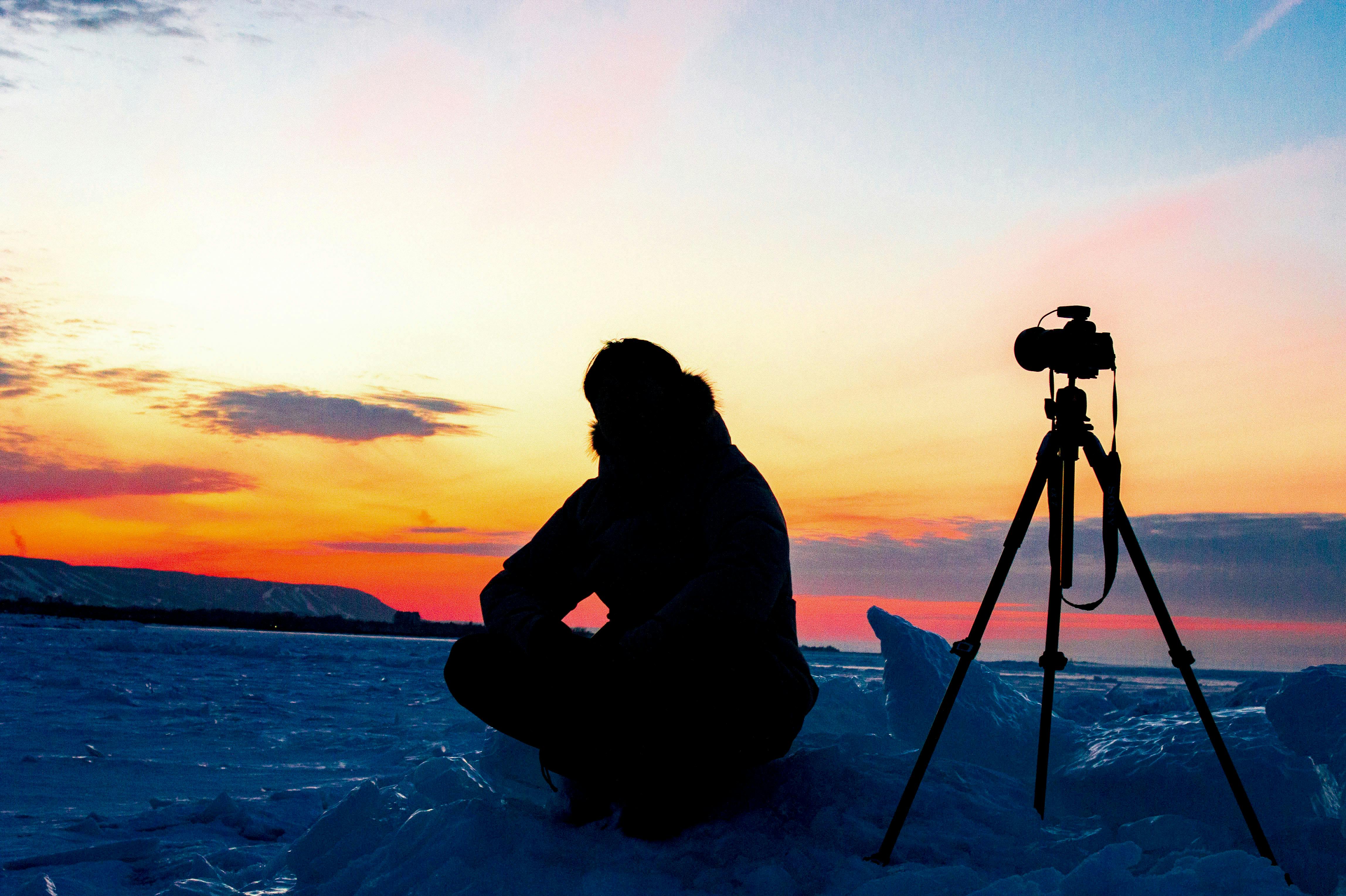 Безкоштовне Фотограф сидить поруч із камерою на штативі на тлі яскравого крижаного заходу сонця. стокове фото