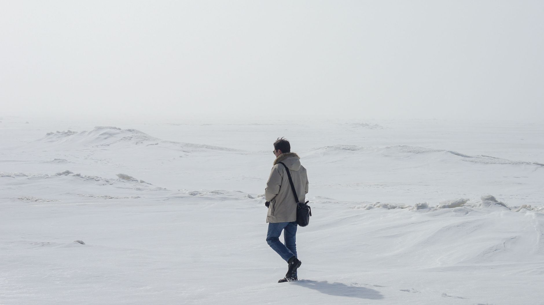 A person in winter clothing walks alone across an expansive snowy field.