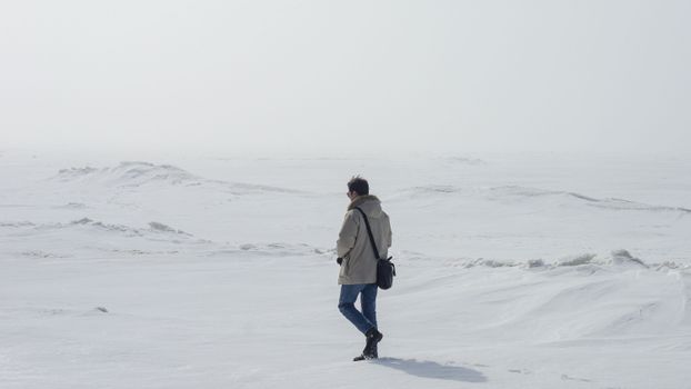 A person in winter clothing walks alone across an expansive snowy field.