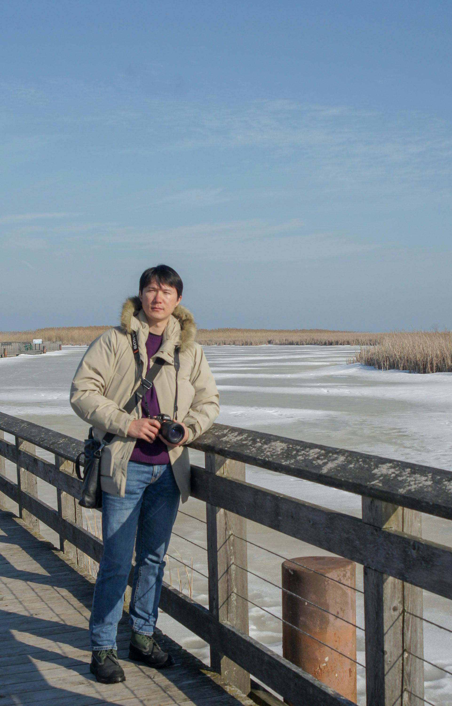 A photographer stands on a boardwalk by a frozen lake under a clear blue sky.