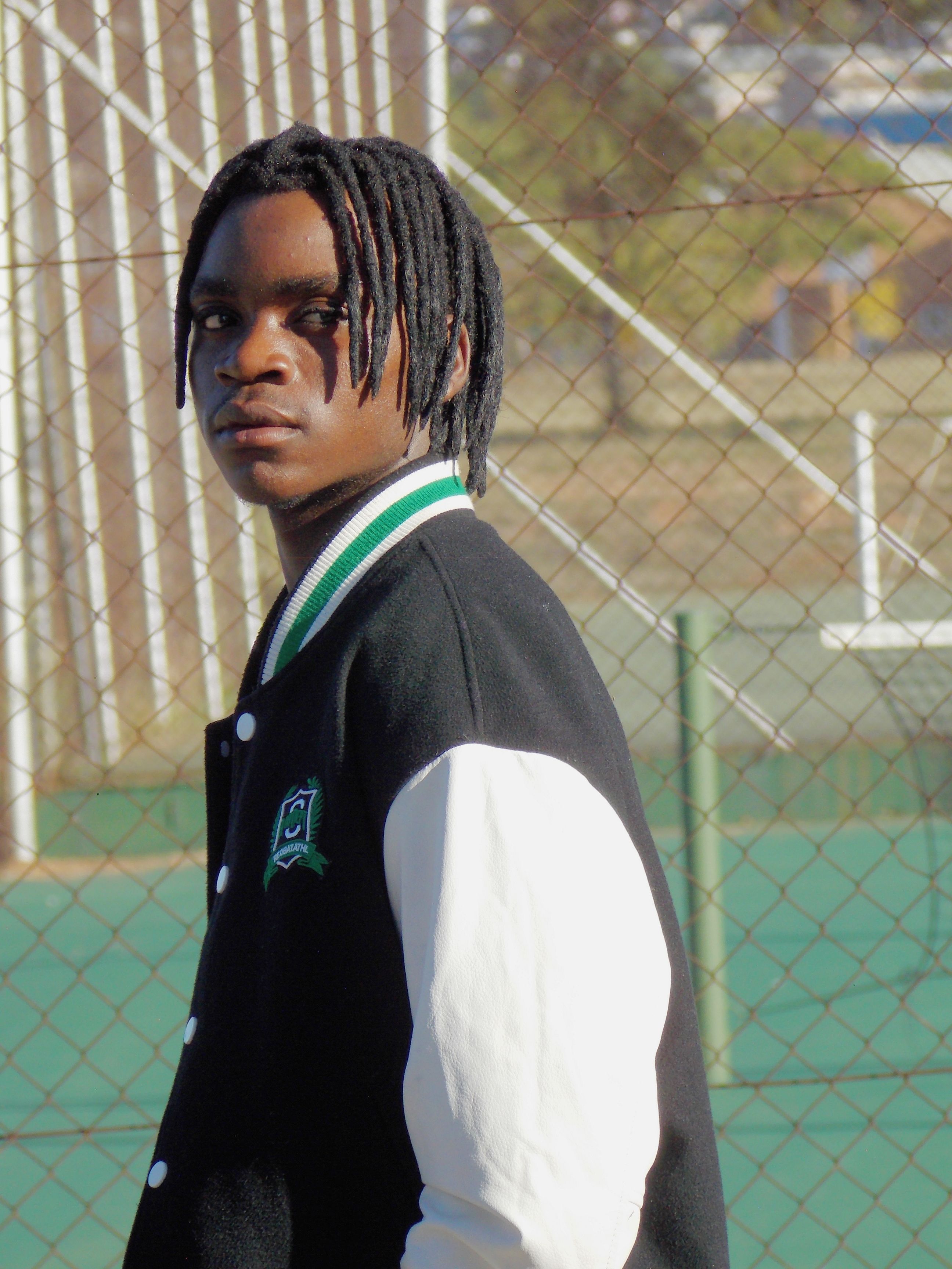 Casual portrait of a young man in a black varsity jacket on a sunlit tennis court.