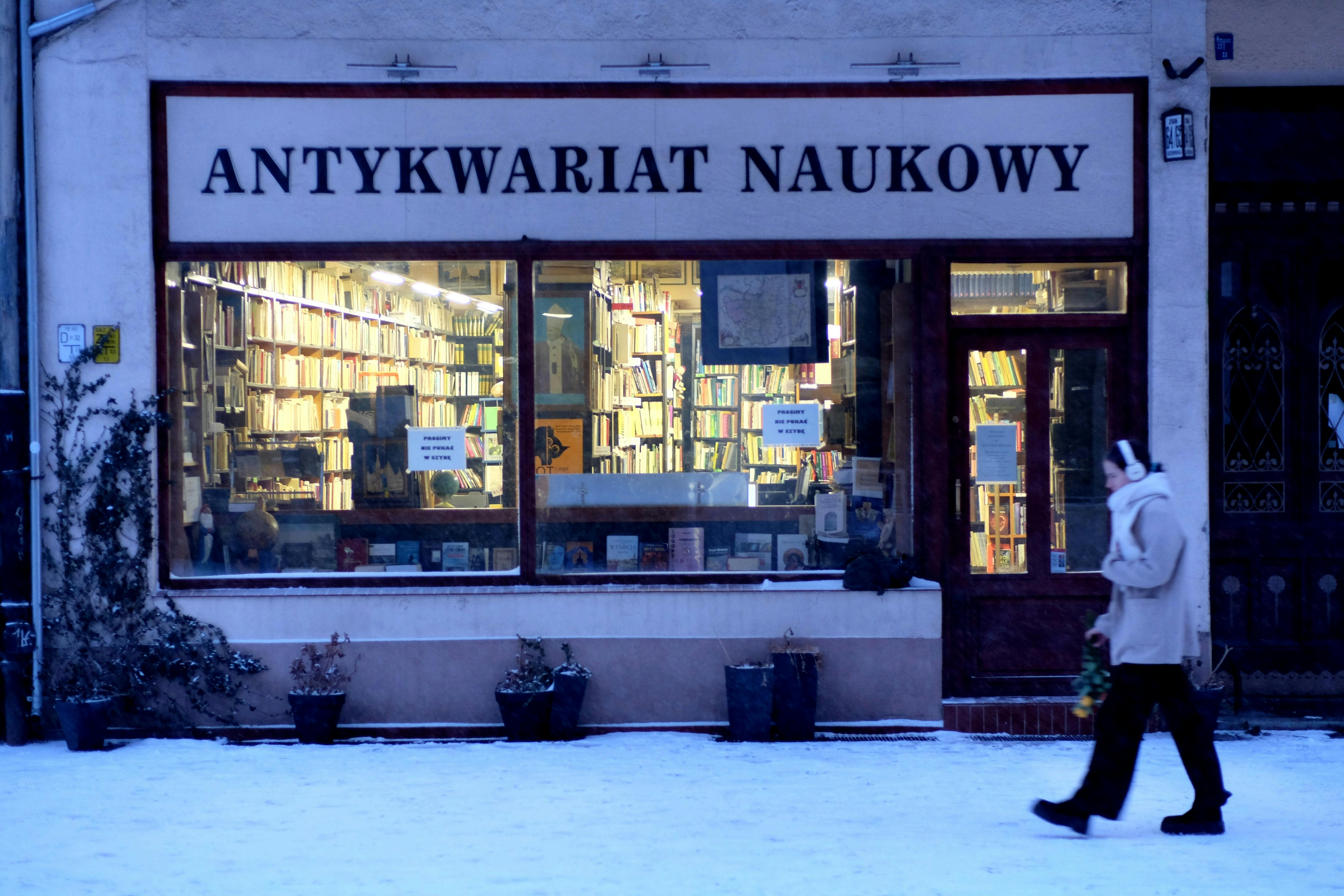 A person walks by a vintage bookstore named 'Antykwariat Naukowy' in a snowy European street.