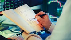 Man Taking Notes with Stock Market Data on Screen