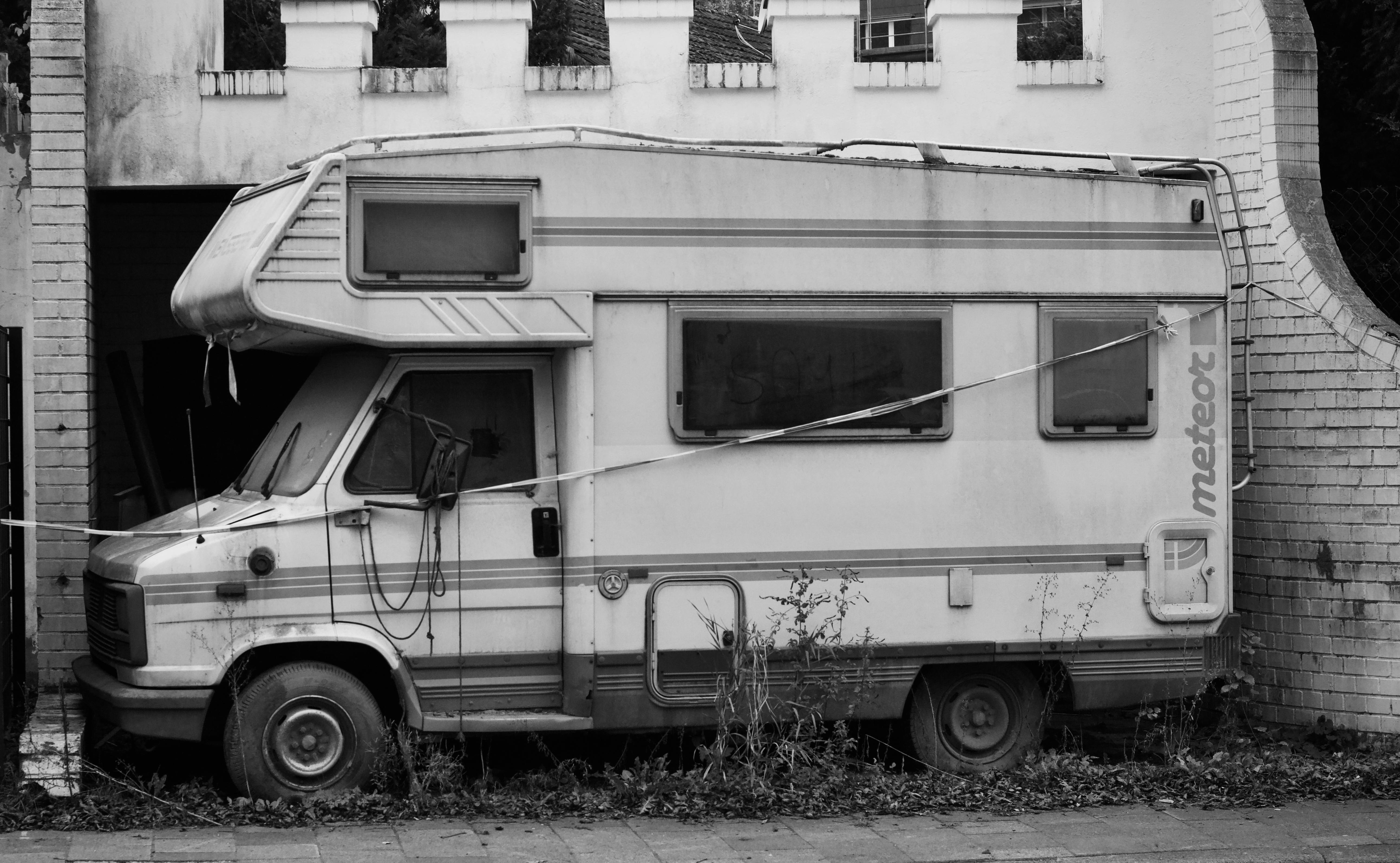 Black and white photo of an old RV parked beside a white brick wall, giving a nostalgic vibe.