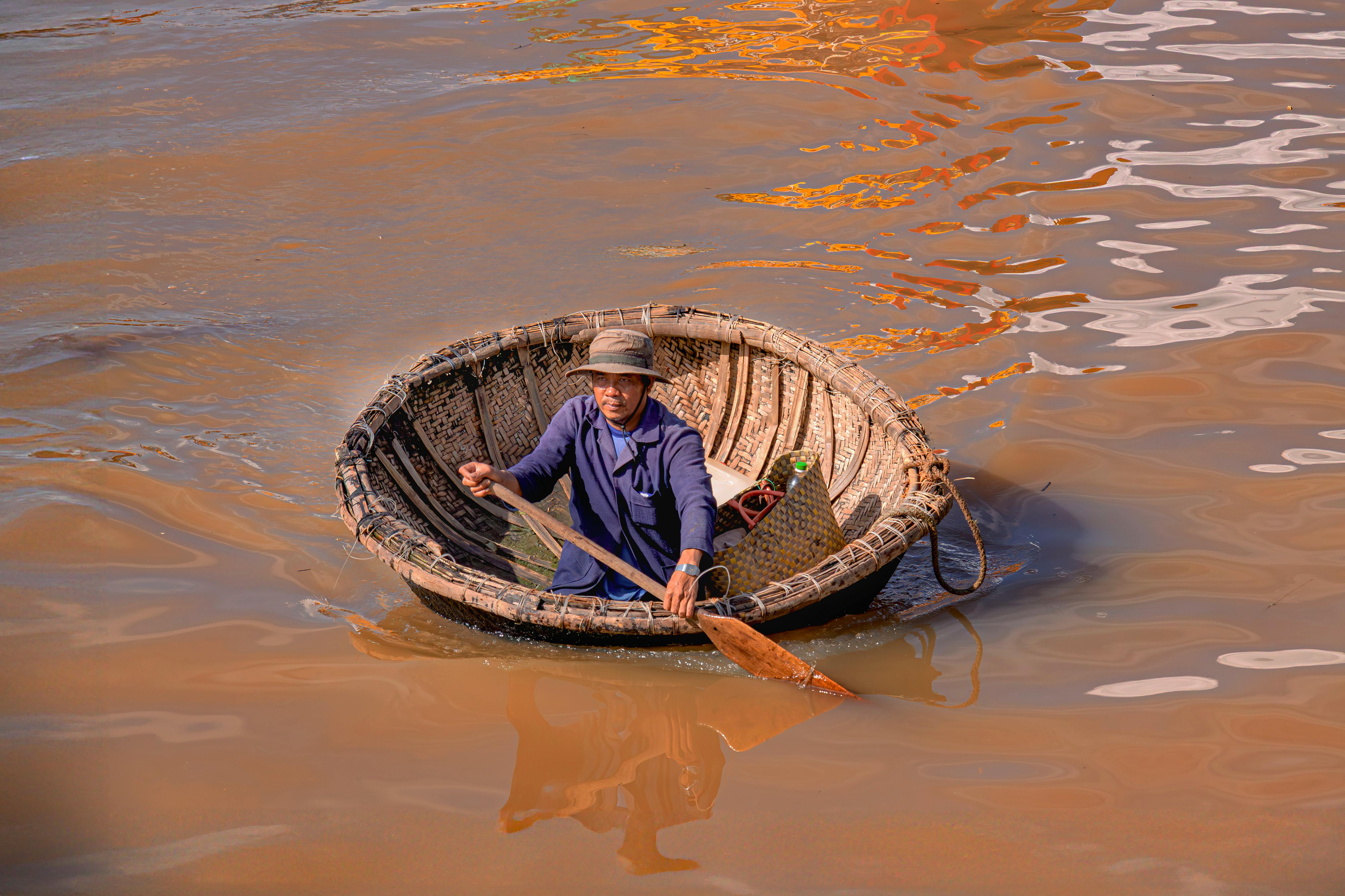 Man in a basket boat rowing on a muddy river in Vietnam, illustrating traditional Vietnamese lifestyle.