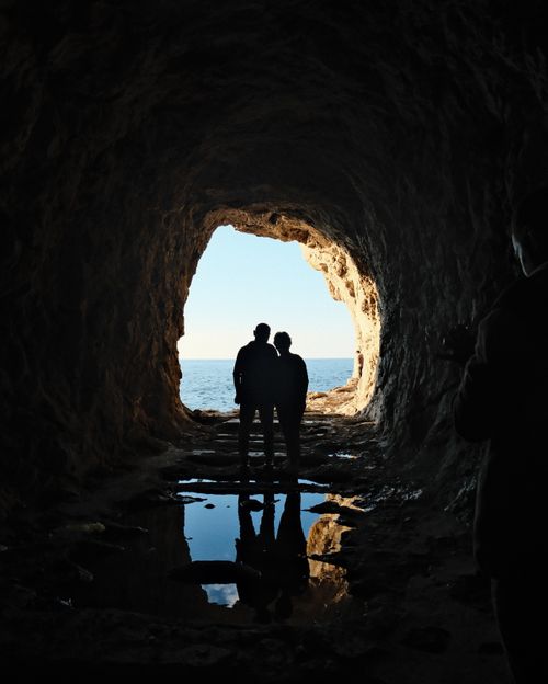 Free Silhouetted couple standing in cave opening, overlooking the sea, creating a dramatic natural frame. Stock Photo