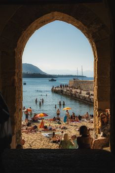 A vibrant beach scene viewed through a stone arch, capturing people enjoying a sunny day by the sea.