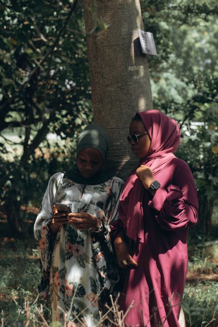 Two women in hijabs enjoy a chat under a tree in a serene outdoor setting.