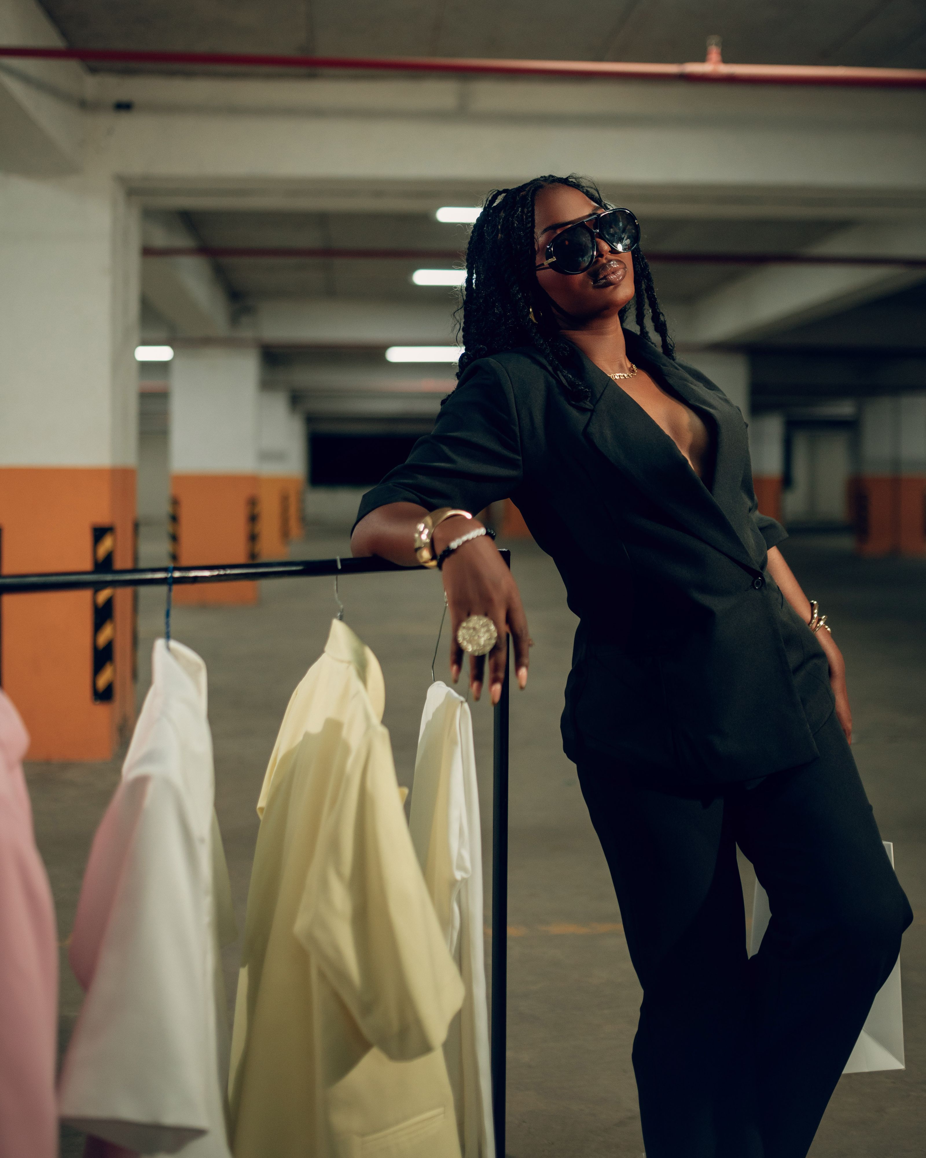Mulher elegante de óculos escuros posando com blazers em um estacionamento urbano sofisticado.