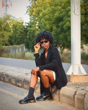Fashionable young woman in sunglasses poses confidently on a city curb in Kano, Nigeria.