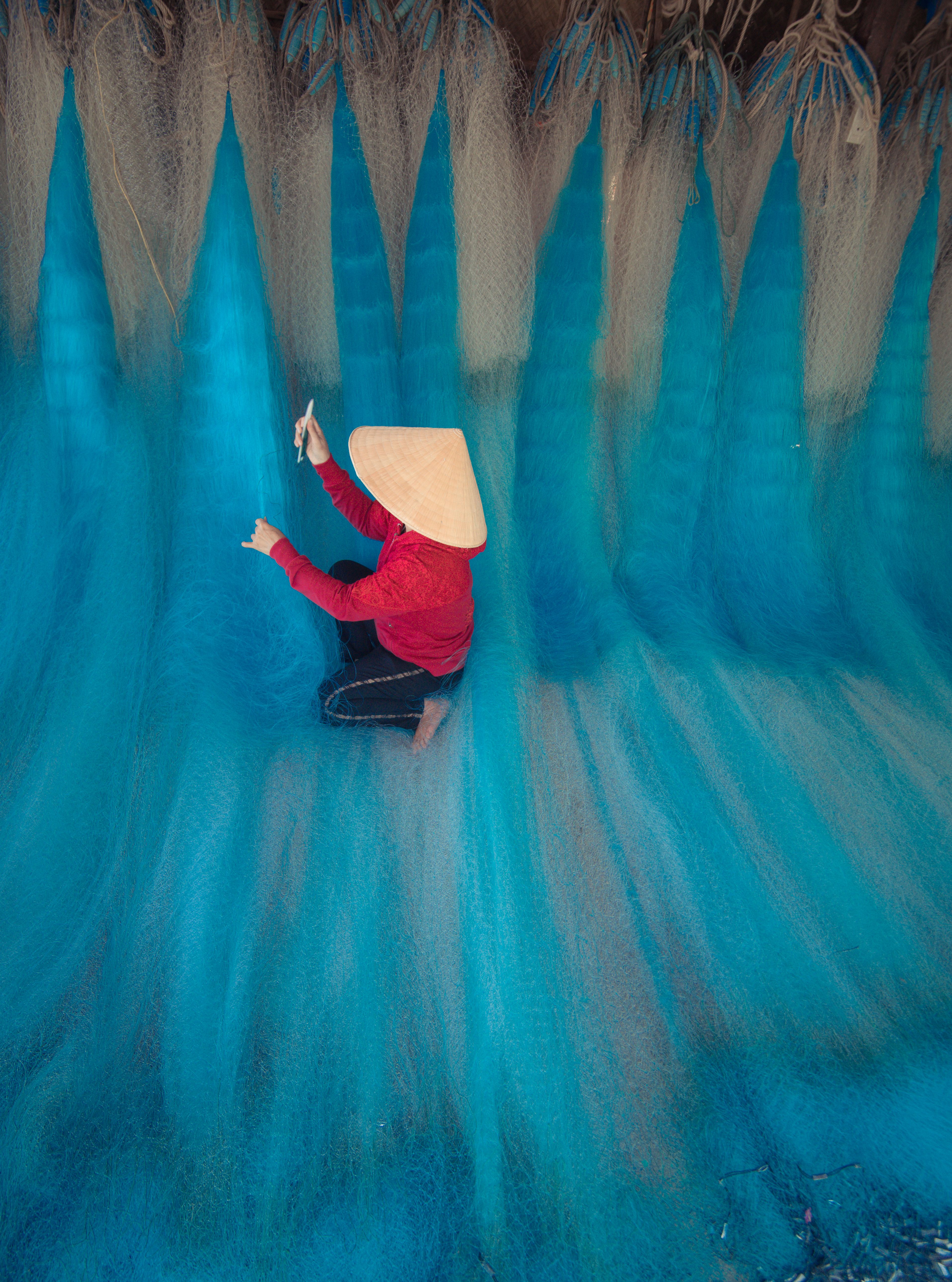 Asian woman in traditional hat weaving vibrant blue fishing nets indoors.