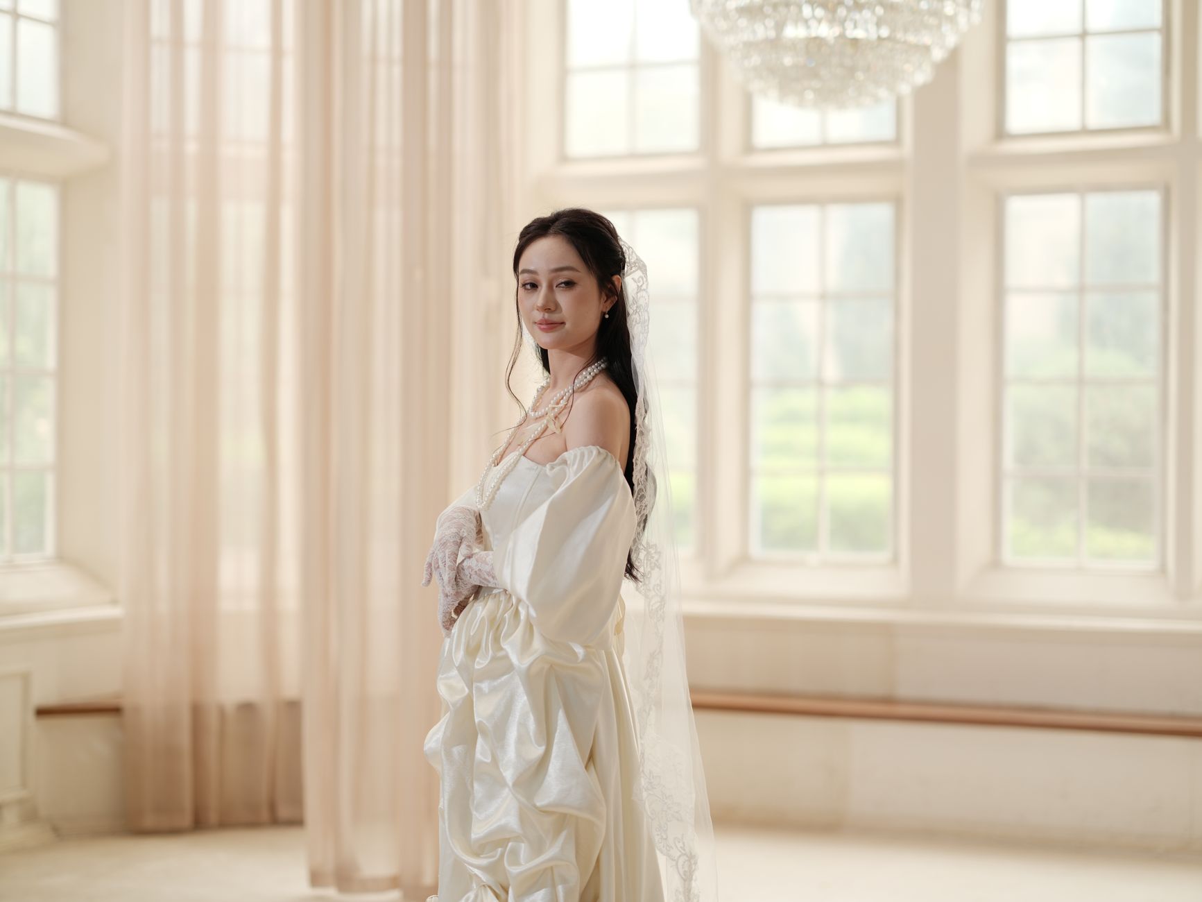 A bride in an elegant gown poses gracefully in a sunlit room with large windows.