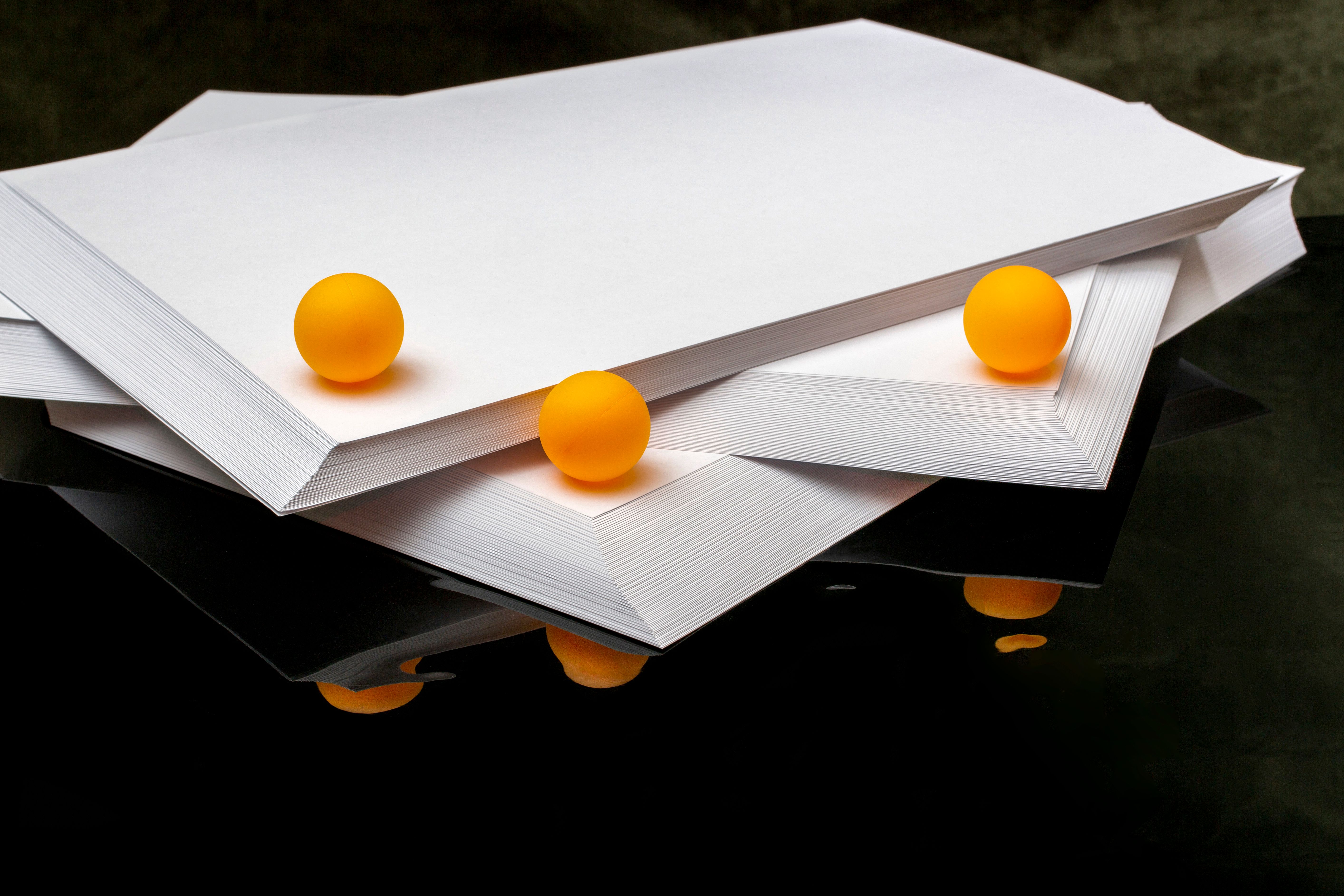 Minimalist composition of paper stacks with vibrant orange balls on reflective surface.