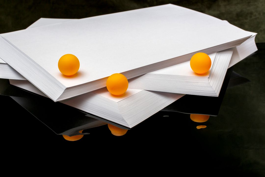 Minimalist composition of paper stacks with vibrant orange balls on reflective surface.