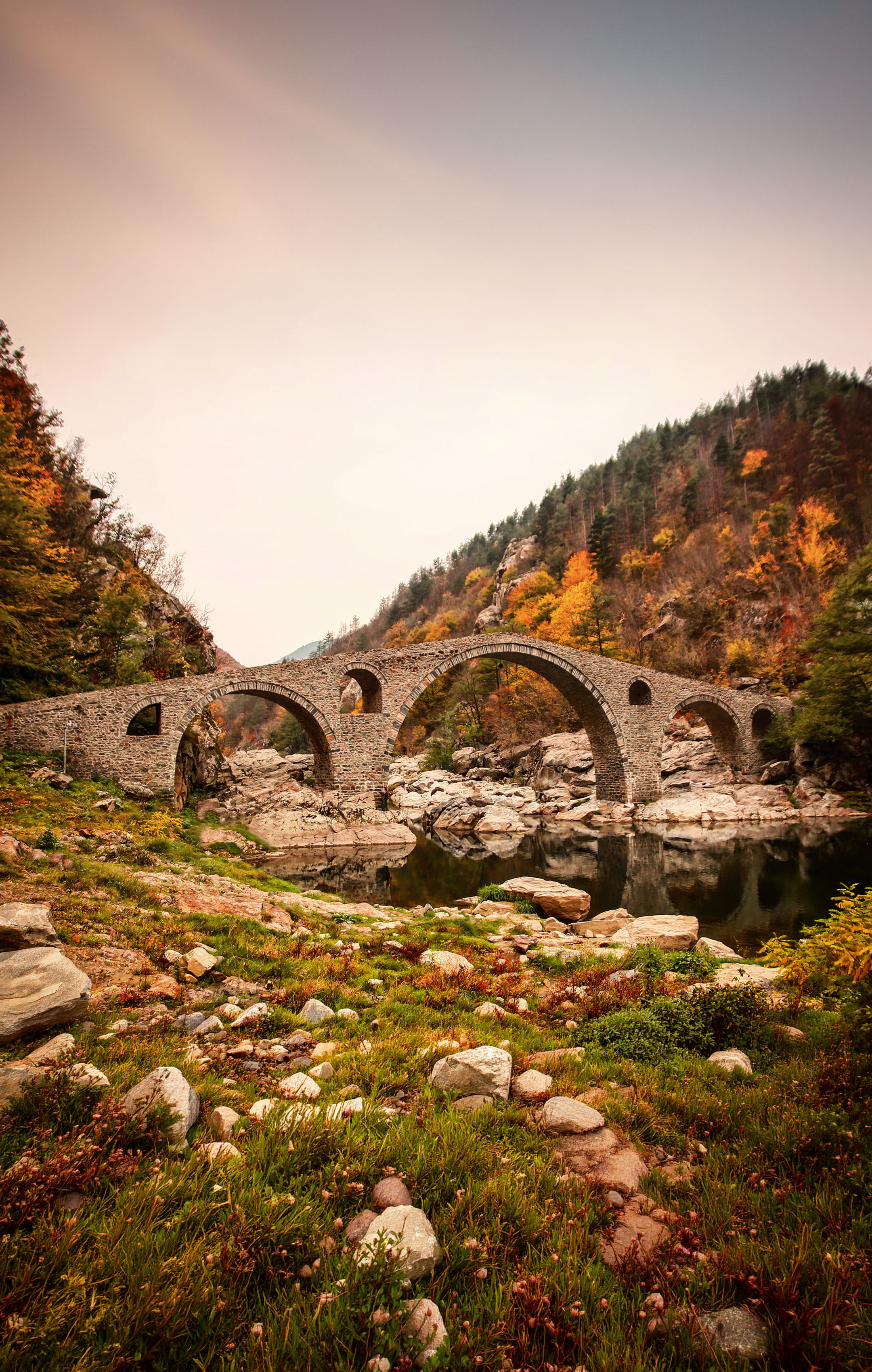 Scenic view of an ancient stone bridge over a river, surrounded by autumn foliage.