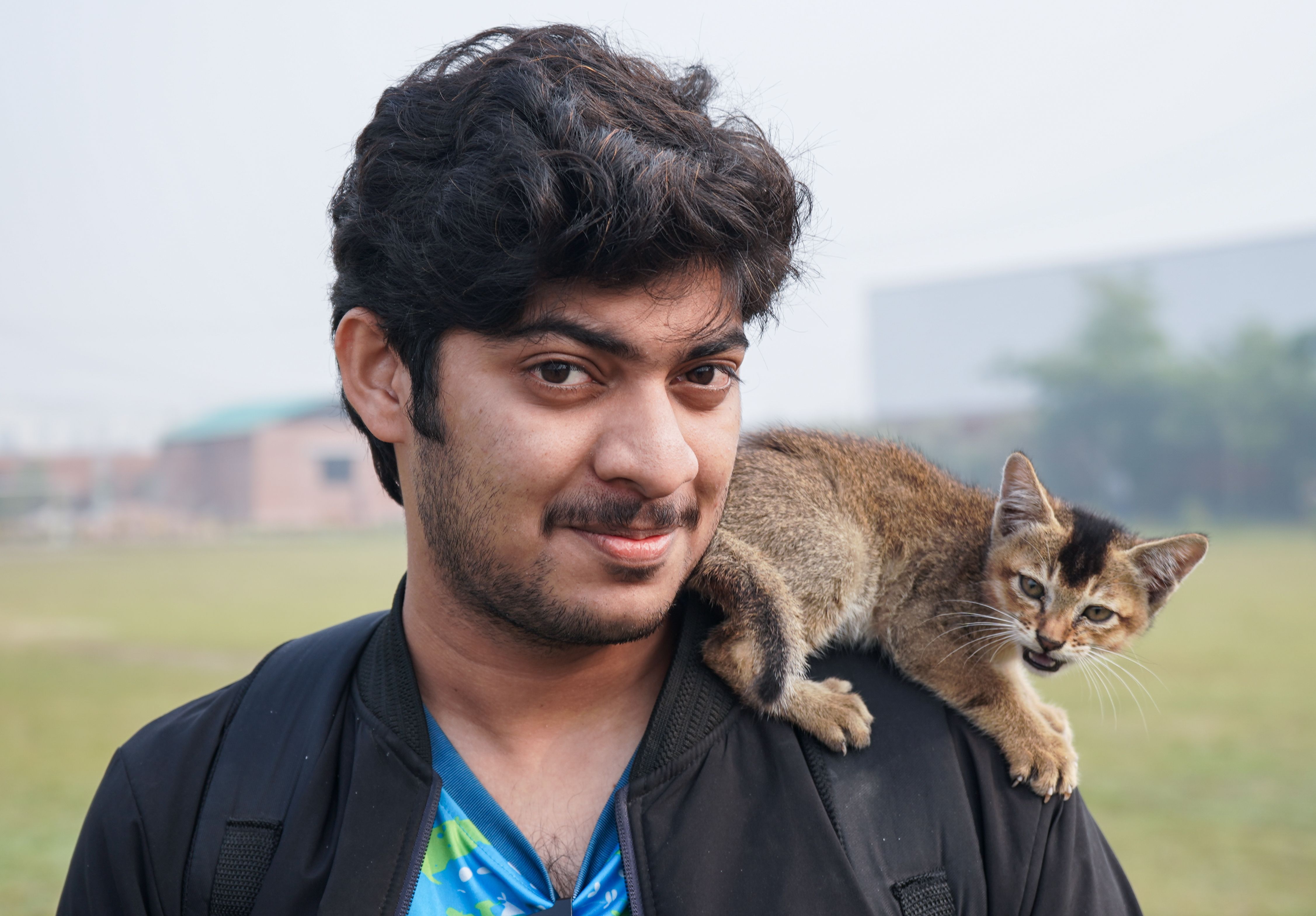 A young man smiles with a cute kitten perched on his shoulder in an outdoor setting in Dhaka, Bangladesh.
