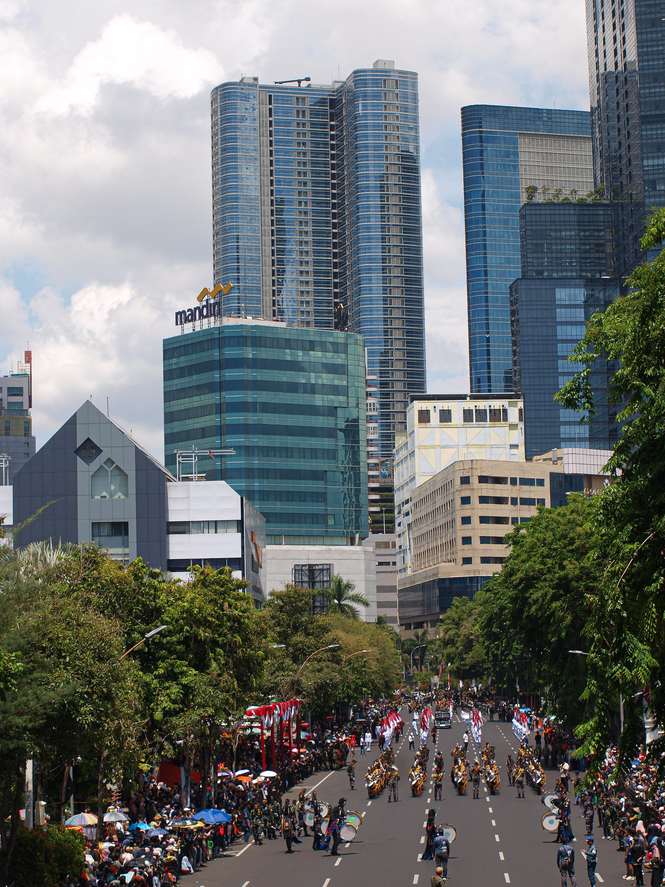 A lively parade unfolds on a sunny day in Jakarta's bustling financial district with tall skyscrapers.