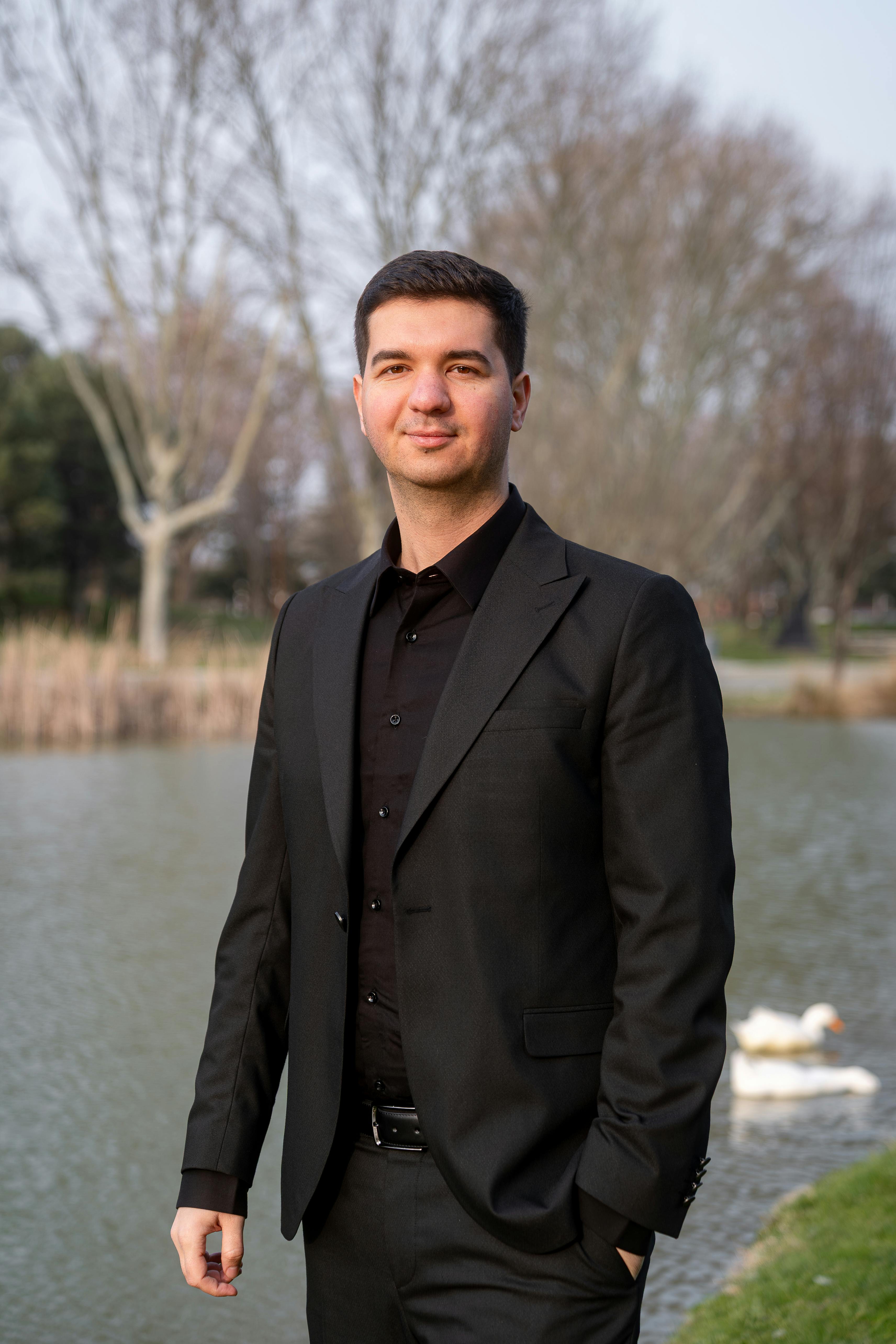 A man in a black suit standing by a riverside, exuding confidence and professionalism.