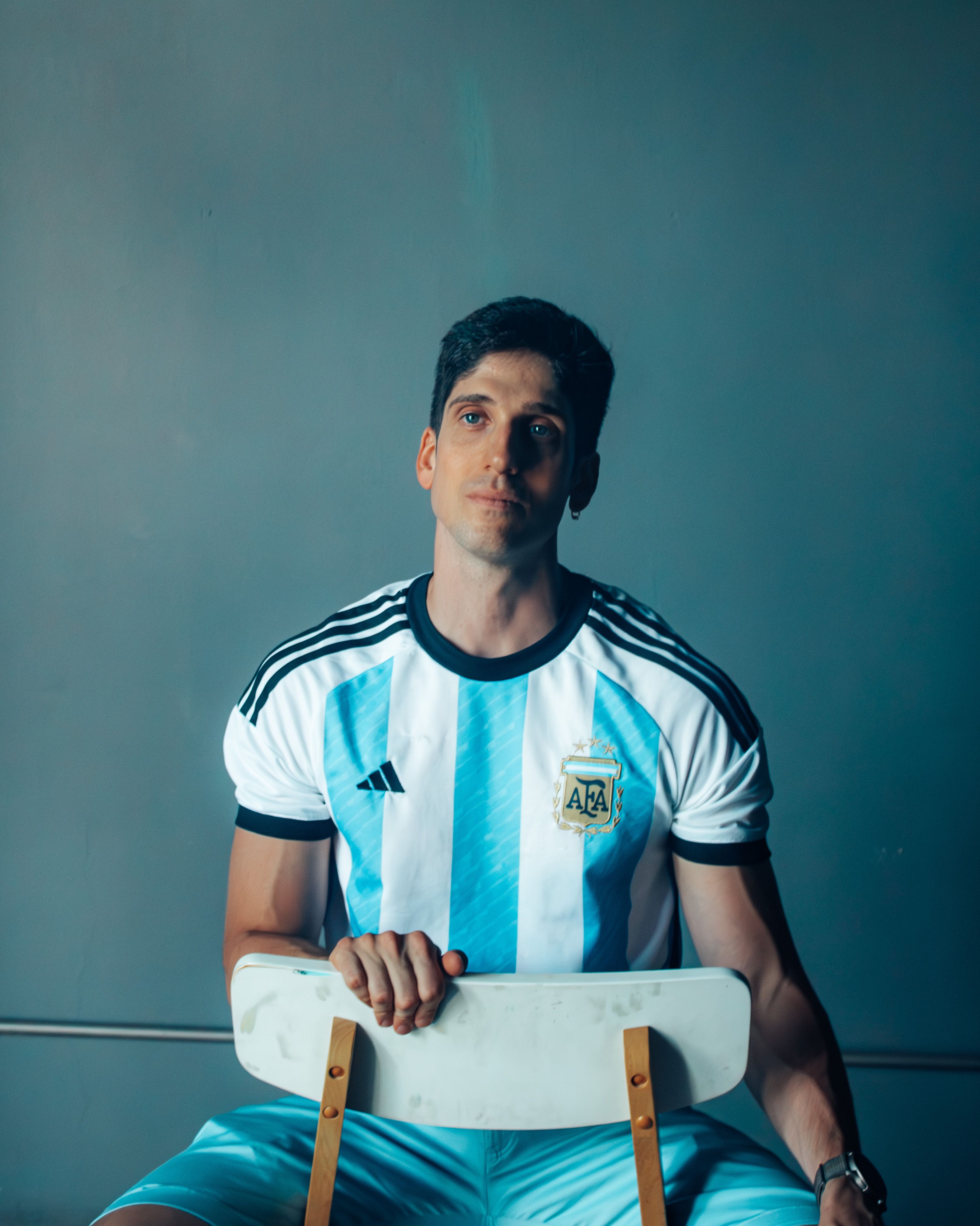 Adult male in Argentina national team jersey seated indoors, gazing upwards.
