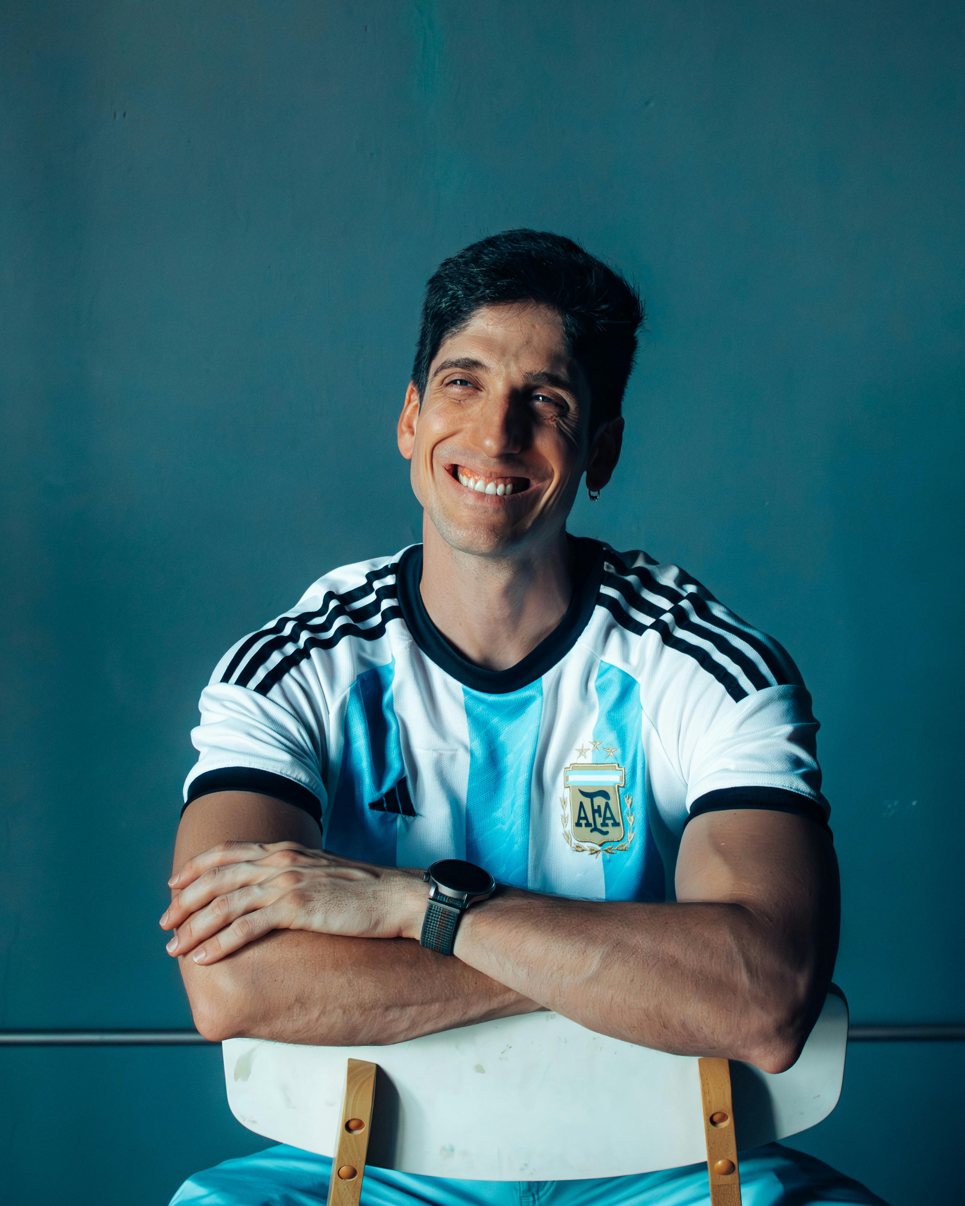 Smiling man wearing an Argentina football jersey seated with arms crossed over a chair.