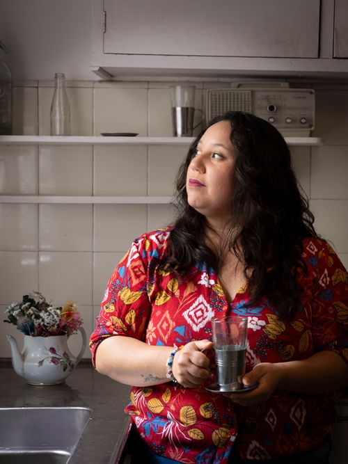 Free A woman in a colorful blouse holds a coffee cup, standing in a cozy kitchen with vintage decor. Stock Photo